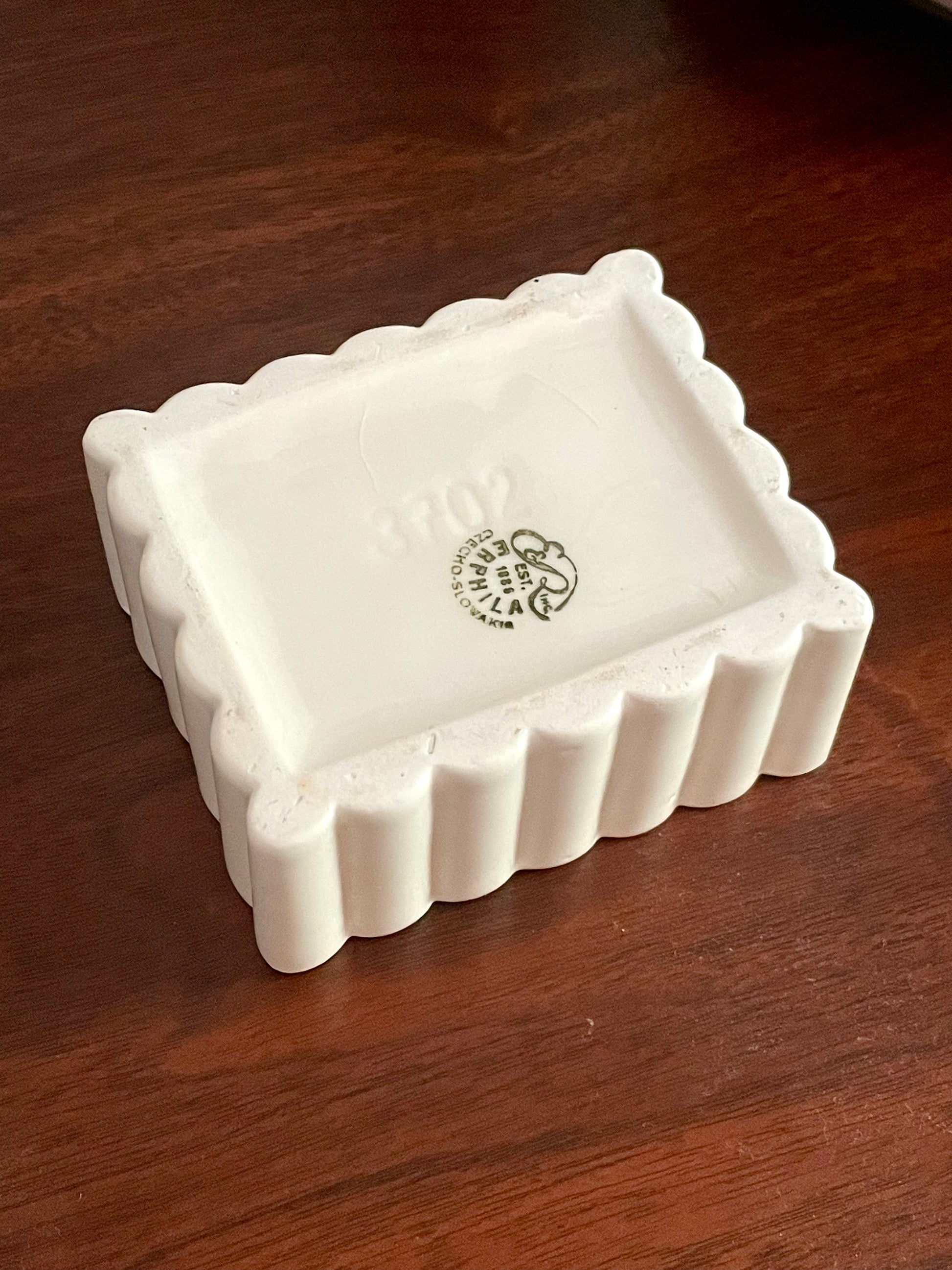 View of the underside of a white ceramic jewelry box with a scalloped edge, photographed on a dark brown wooden surface. The inside of the tray features a dark green stamp with ‘Erphila Czecho-Slovakia'.