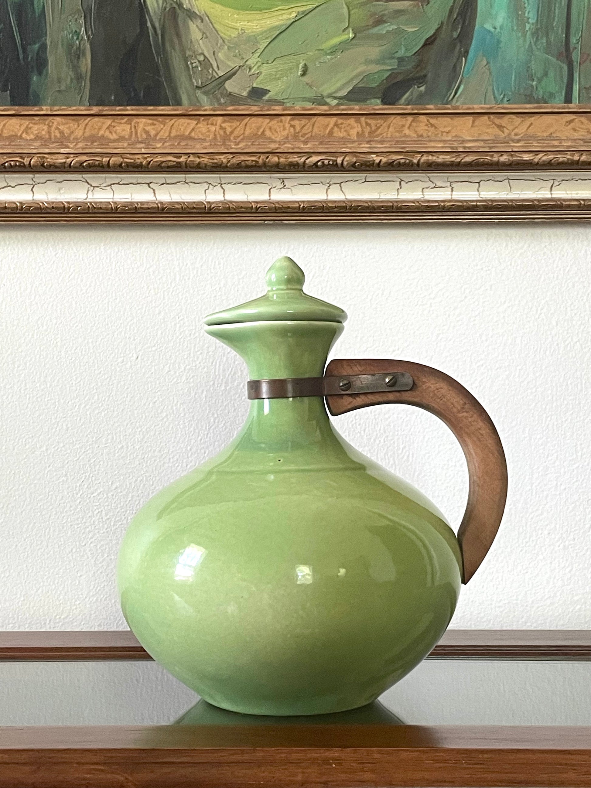A light green ceramic pitcher with a lid and a curved brown wooden handle, displayed on a wooden surface against a white wall with a framed painting in the background.