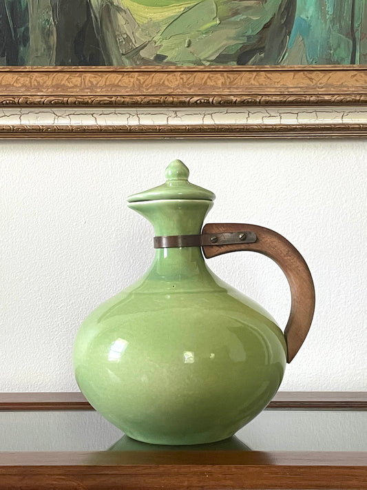 A light green ceramic pitcher with a lid and a curved brown wooden handle, displayed on a wooden surface against a white wall with a framed painting in the background.