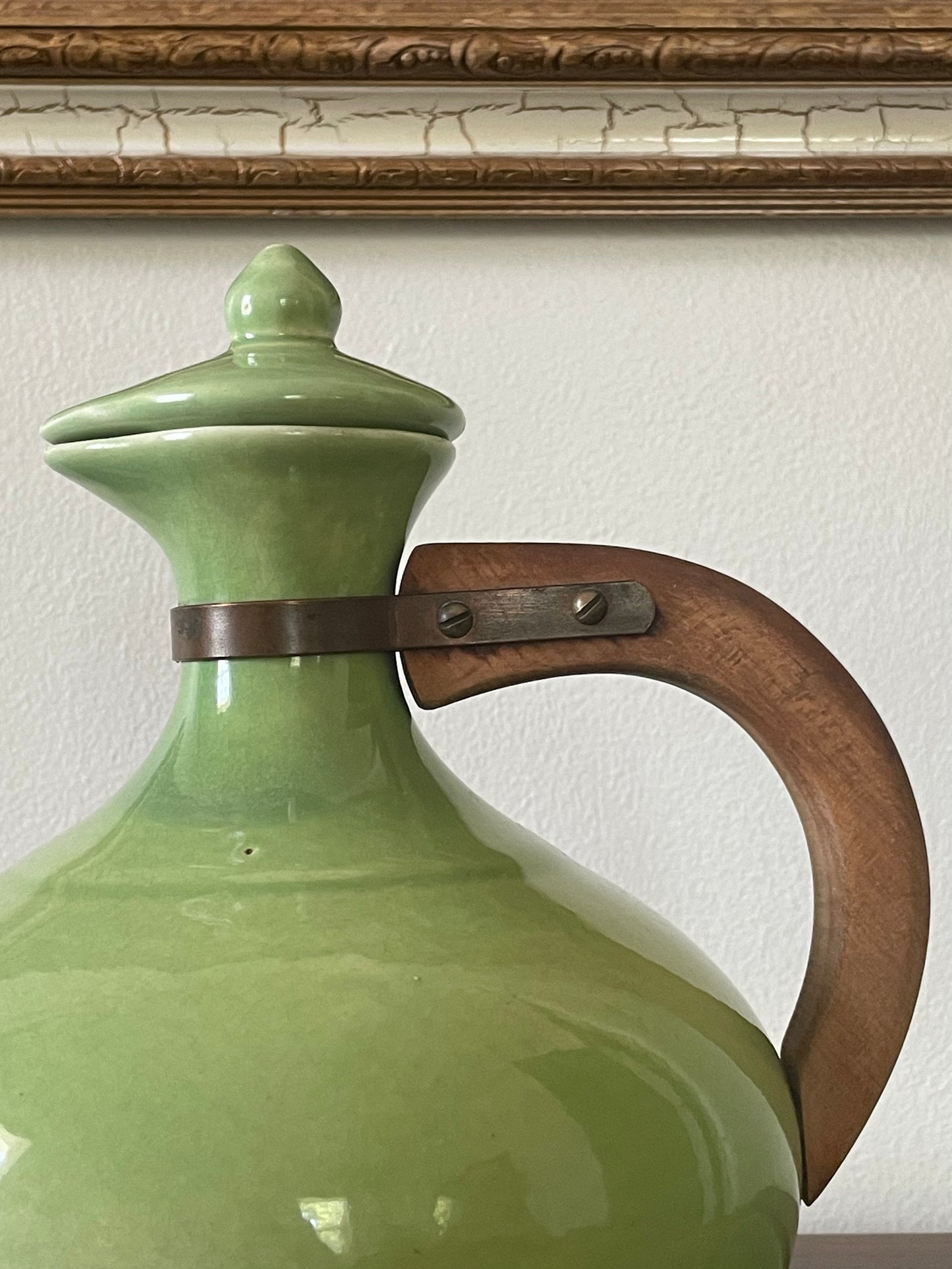 Close-up of a glossy light green ceramic carafe with a matching lid, featuring a curved brown wooden handle secured by a dark metal band, set against a white wall and wood trim.