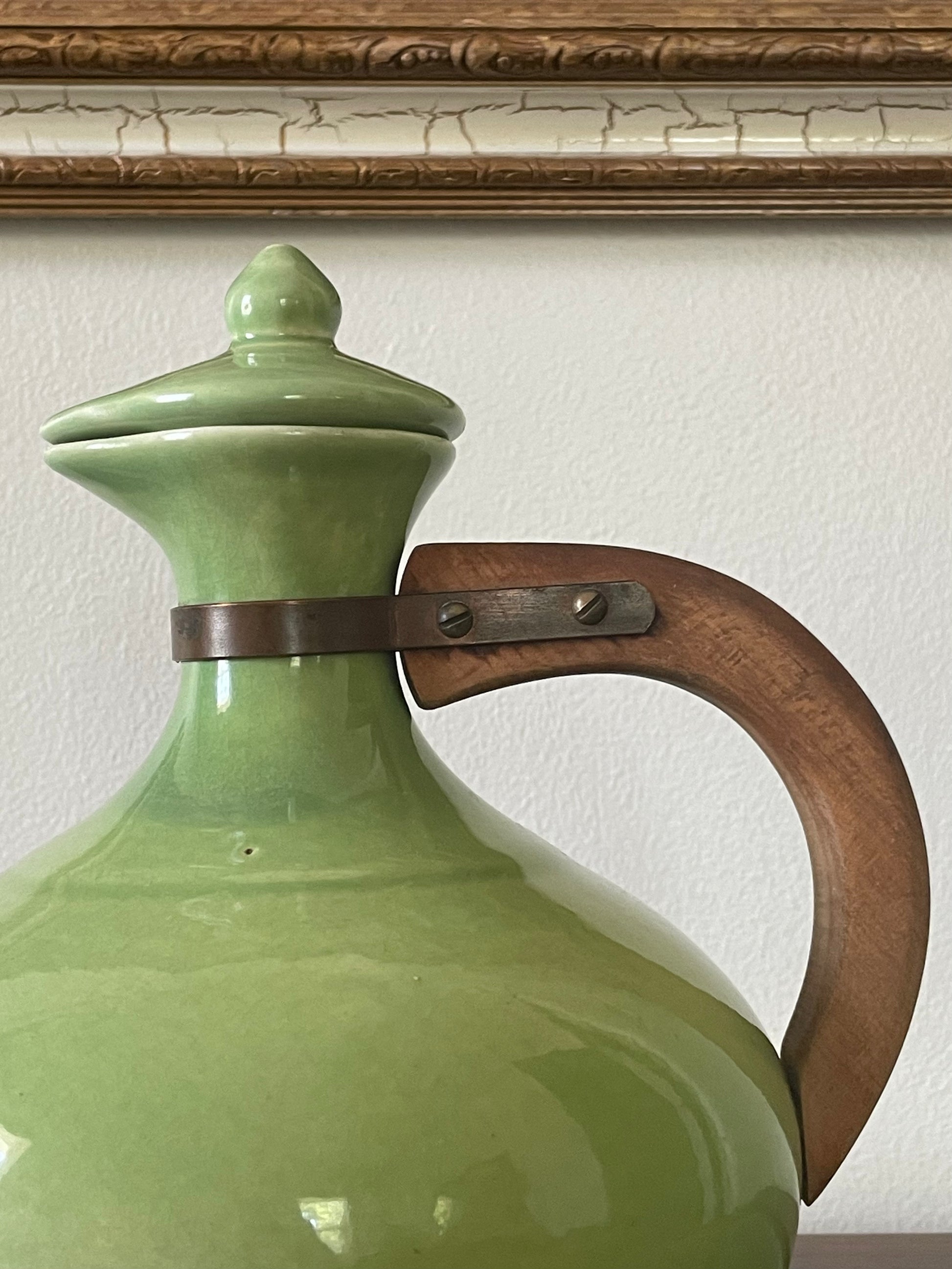 Close-up of a glossy light green ceramic carafe with a matching lid, featuring a curved brown wooden handle secured by a dark metal band, set against a white wall and wood trim.