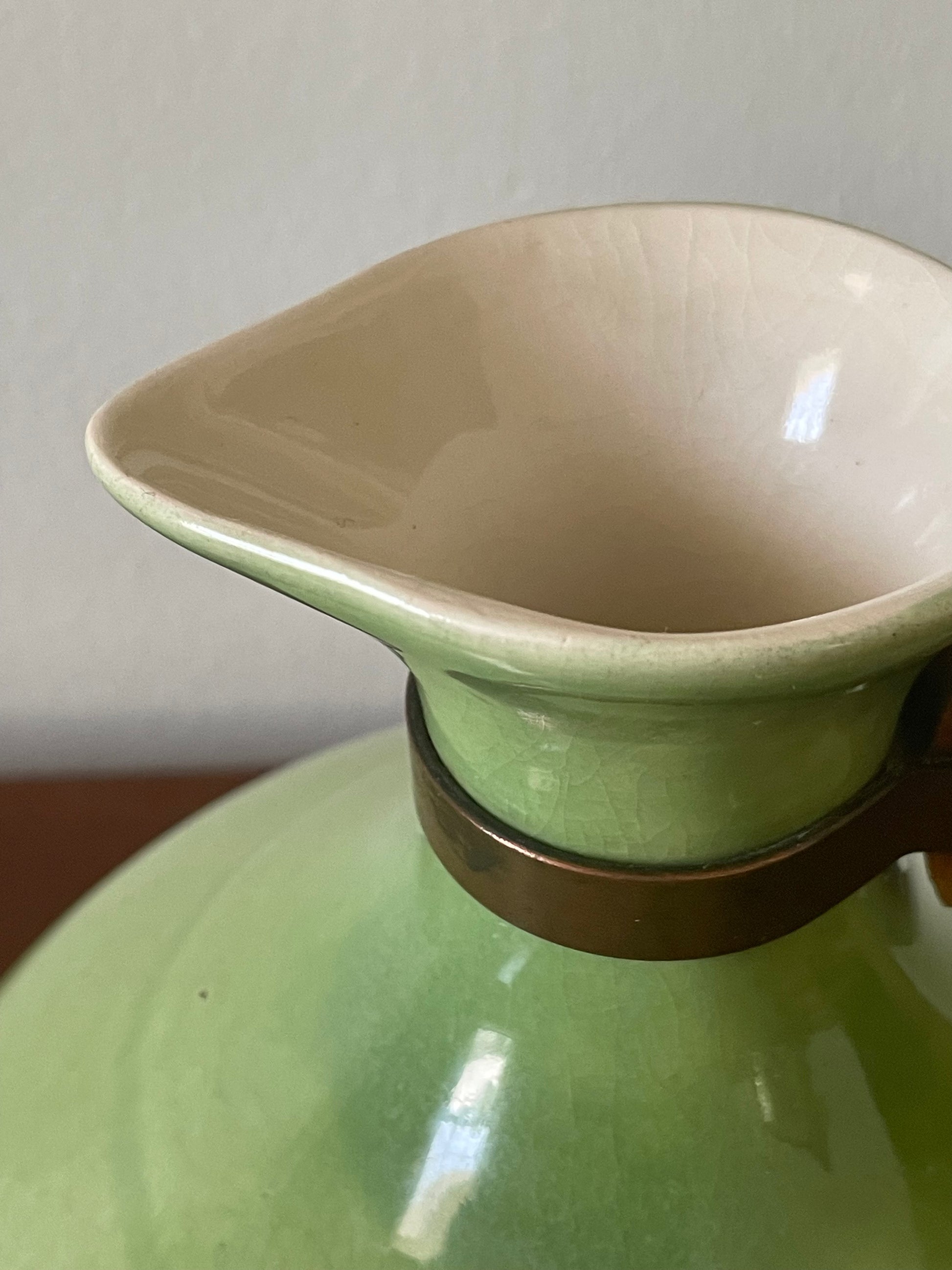 Close-up of a ceramic carafe with a light green exterior, a cream-colored interior, and a dark metallic band around the neck.