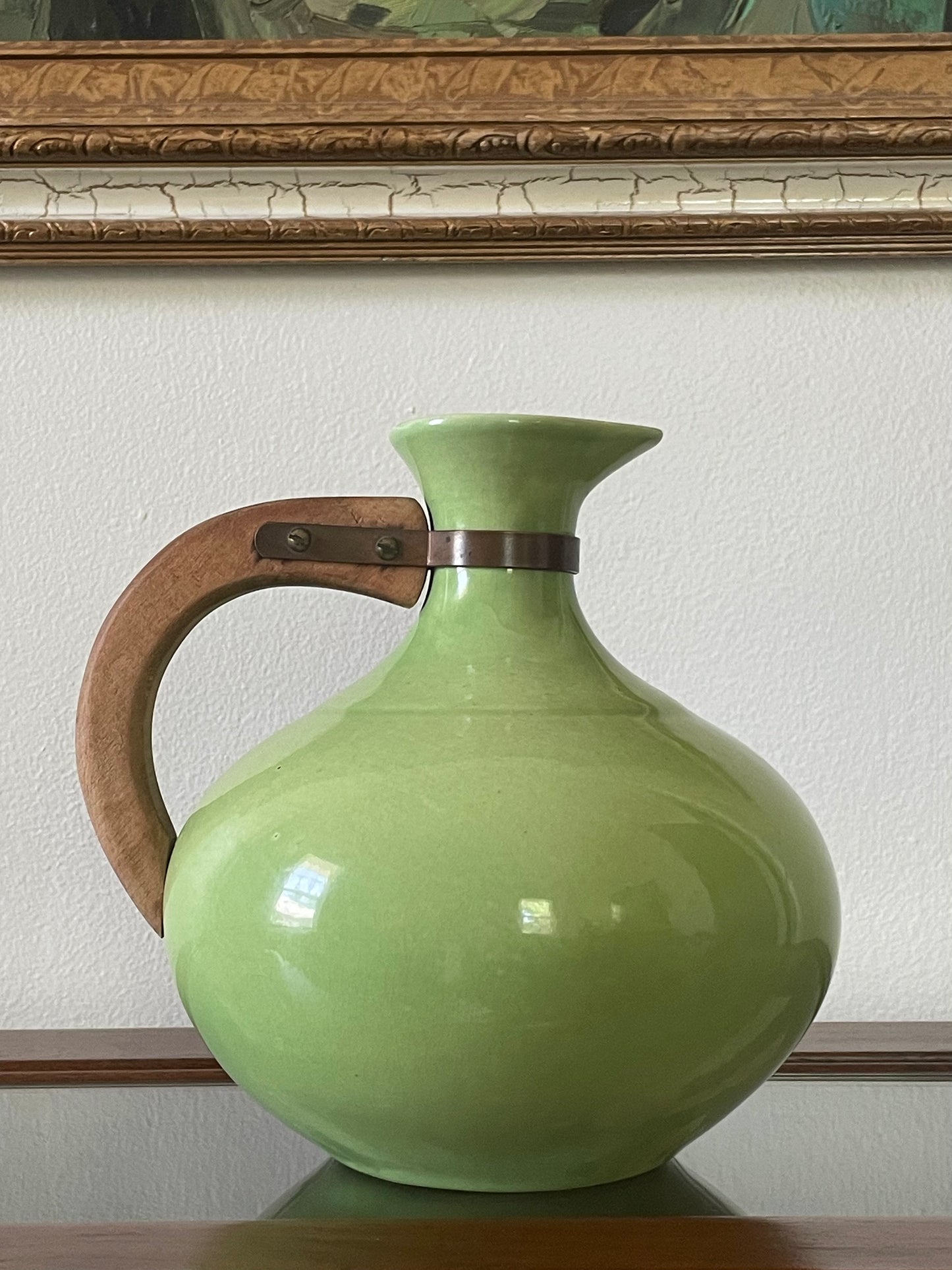 A light green ceramic pitcher with a wooden handle and a dark metal collar, displayed on a wooden surface against a white wall with a picture frame visible above.