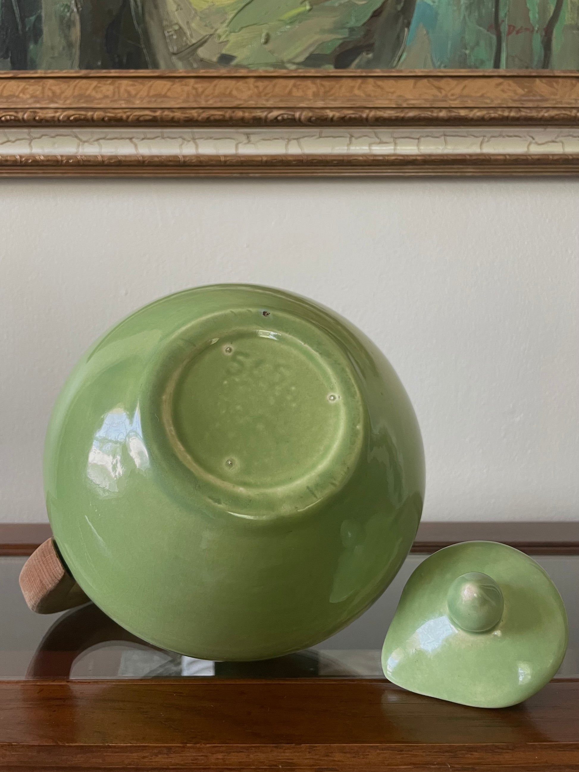 Underside of a glossy light green ceramic carafe with ‘565’ stamped on the bottom with its matching lid placed beside it on a wooden surface, with a cream wall and framed artwork in the background.