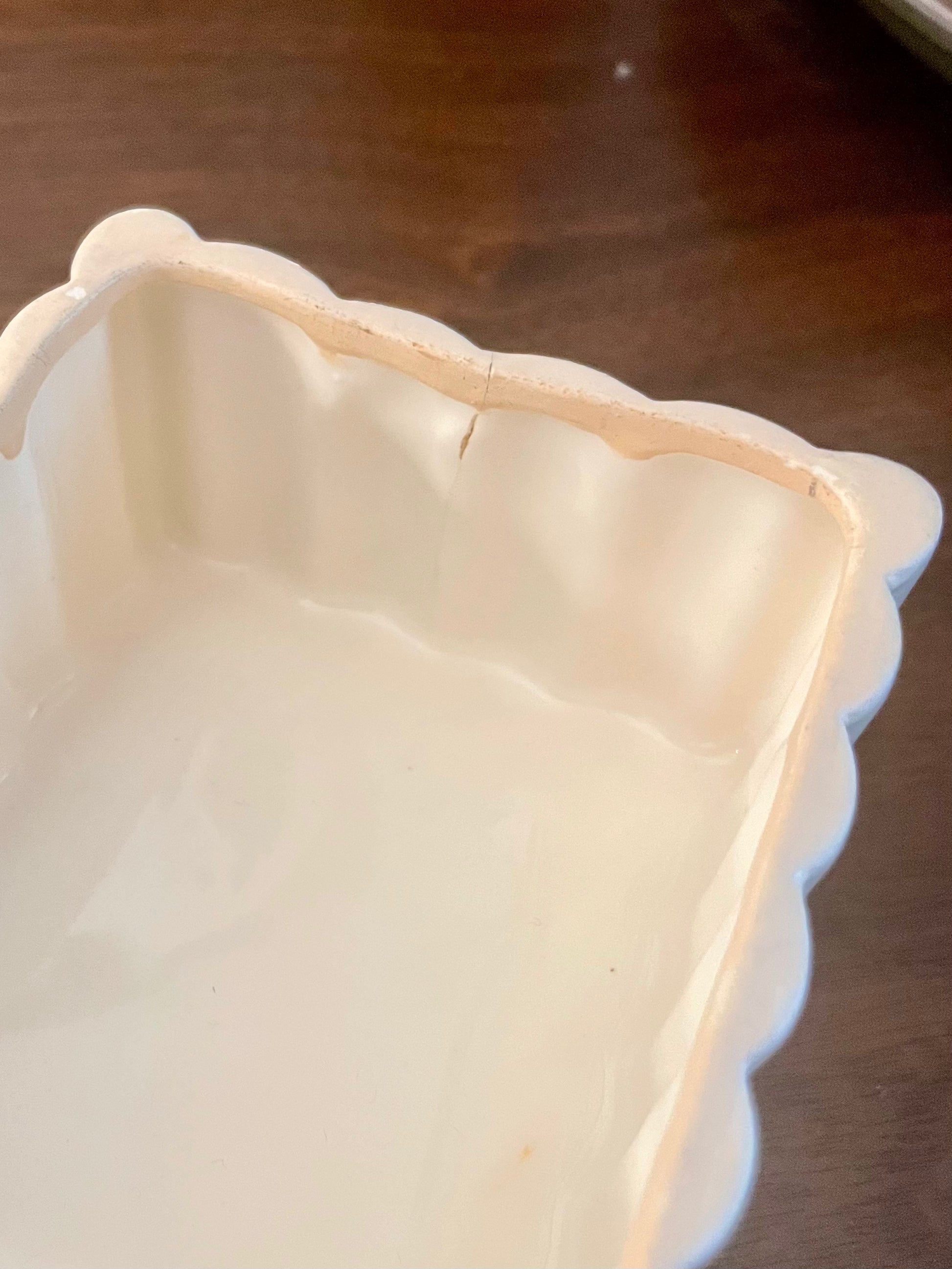 Closeup of the inside of an off-white ceramic box with a scalloped rim showing a hairline crack, photographed on a dark wooden tabletop.