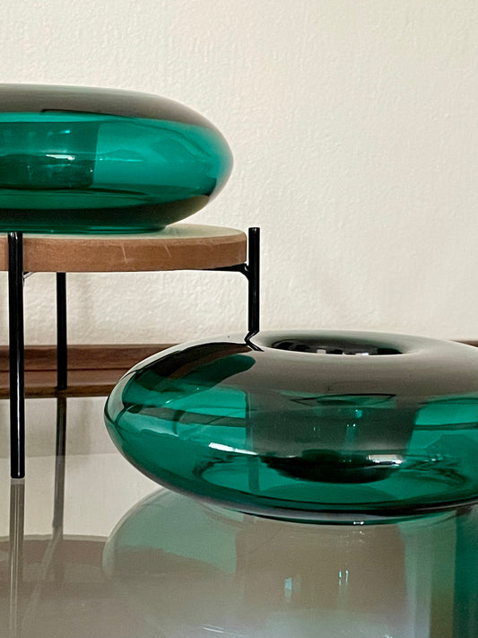 Two translucent emerald green glass vessels that are donut-shaped with a central indent to hold votive candles, displayed on a glass table. One of the candleholders rests on a wood and metal riser.