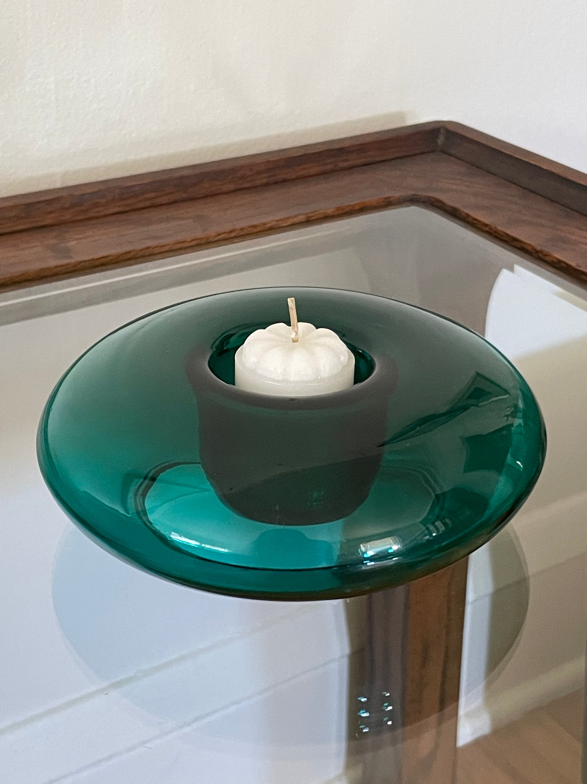 A round translucent emerald green glass candle holder with a central indent holding a small white candle, set on a glass-topped table with a wooden frame.