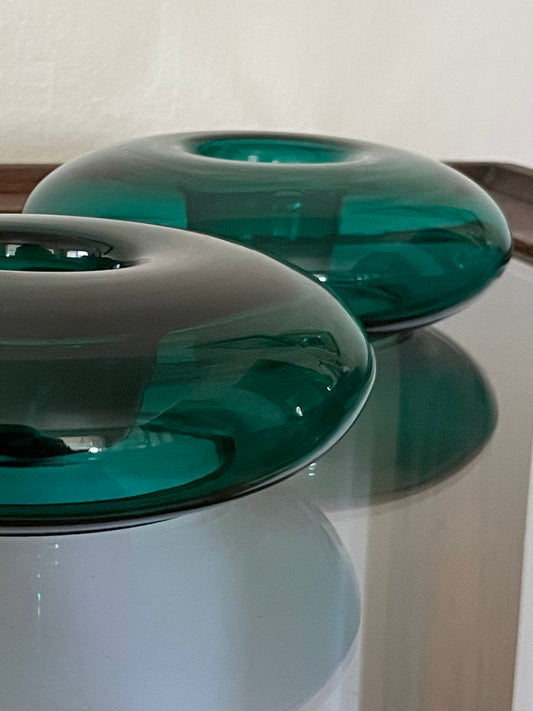 Two translucent dark green glass candle votive holders with a smooth, glossy finish, displayed on a reflective glass surface against a neutral light background.