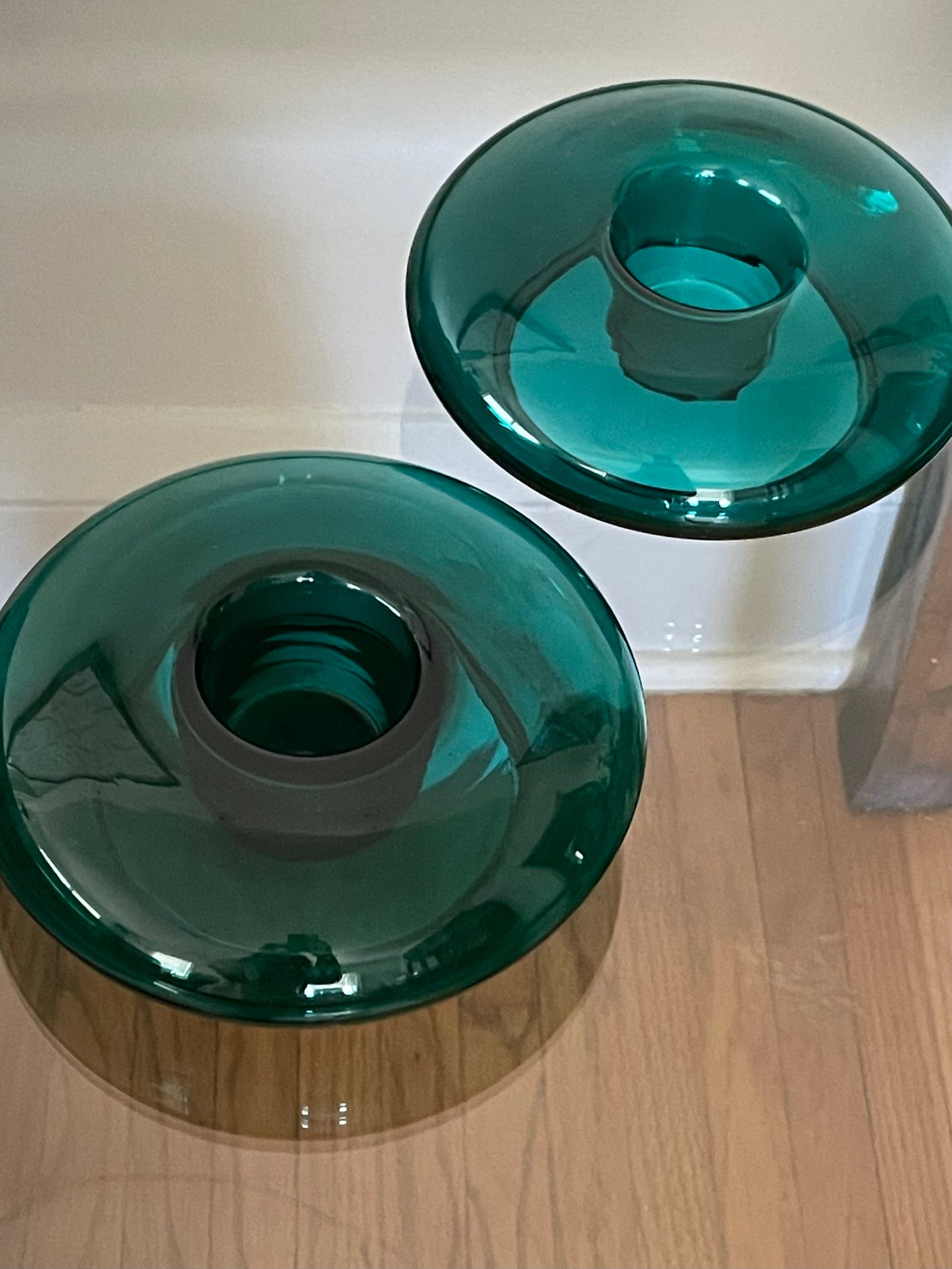 Two translucent dark green glass candle holders with a donut shape and central indent, displayed on a glass-top wood table against a white wall.