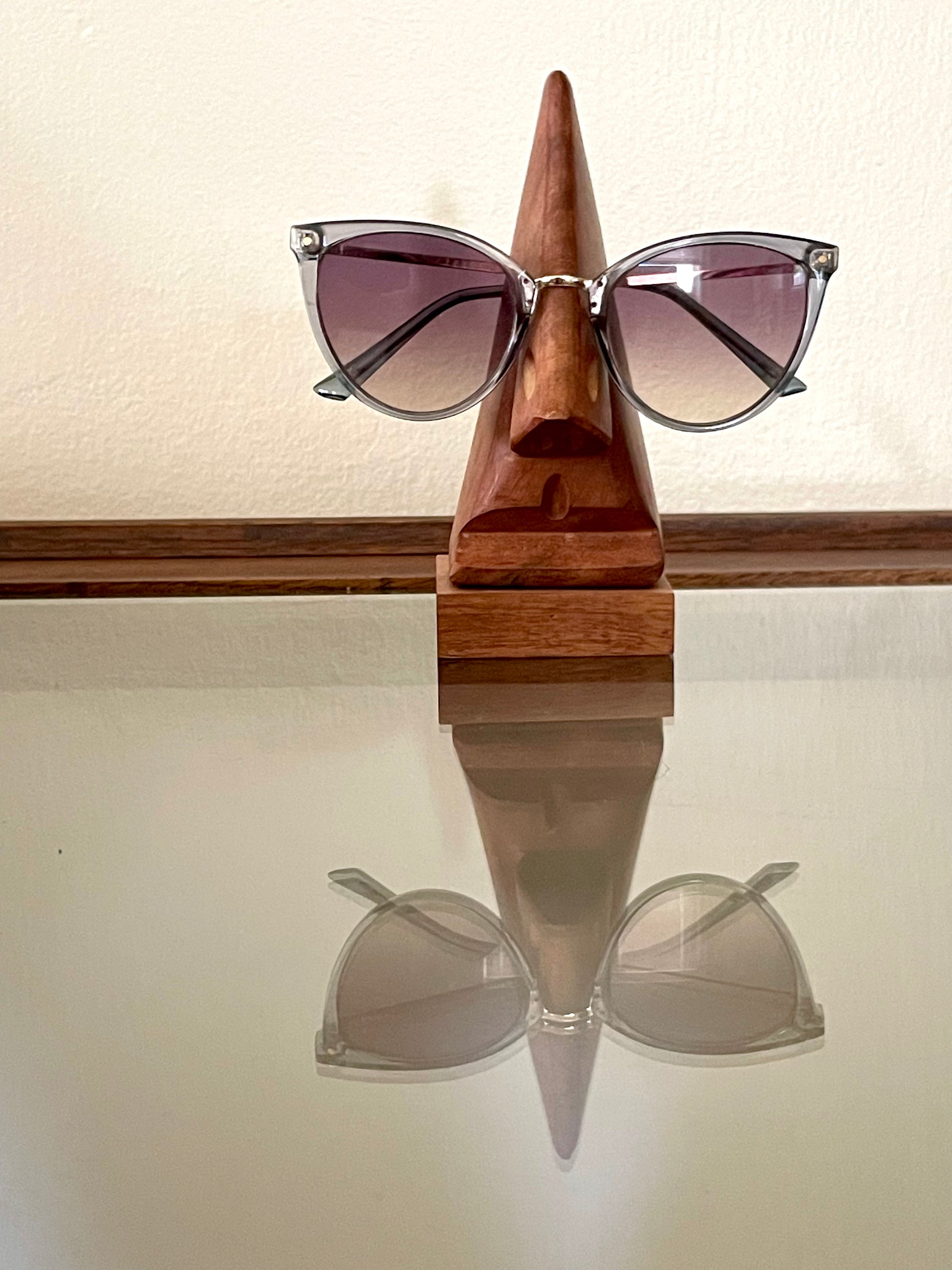 Cat-eye sunglasses with a gradient purple tint and silver metal frames, displayed on a triangular wooden stand shaped like a stylized face against a beige wall.