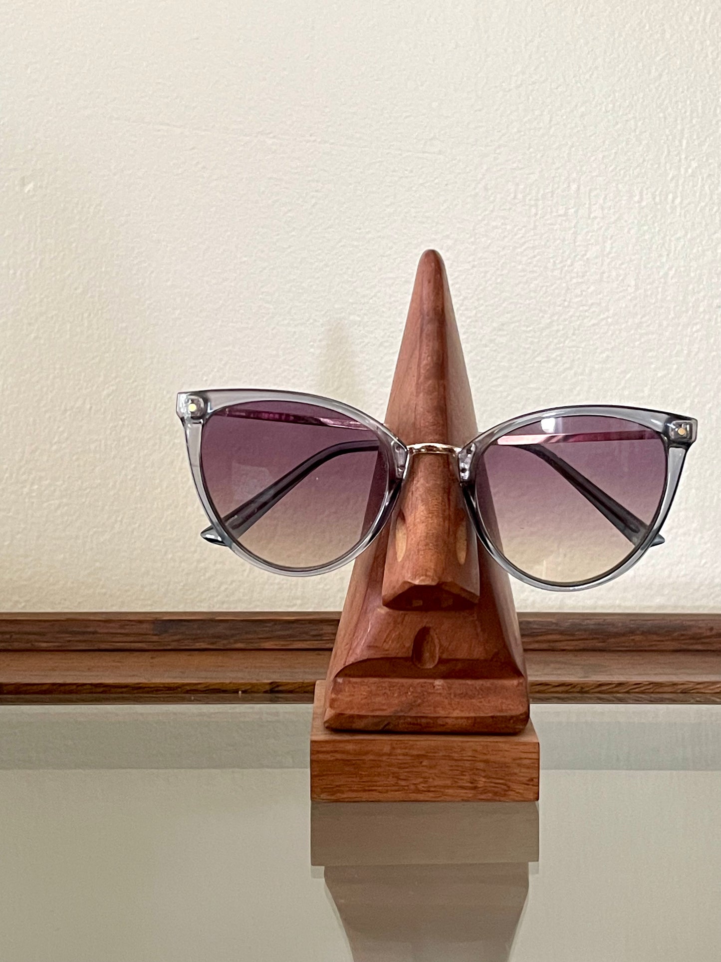 Cat-eye sunglasses with gradient purple lenses and silver metal frames, displayed on a triangular wooden stand carved in the likeness of a nose and mouth, set against a cream-colored wall.
