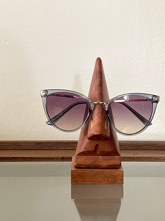 Cat-eye sunglasses with gradient purple lenses and silver metal frames, displayed on a triangular wooden stand carved in the likeness of a nose and mouth, set against a cream-colored wall.