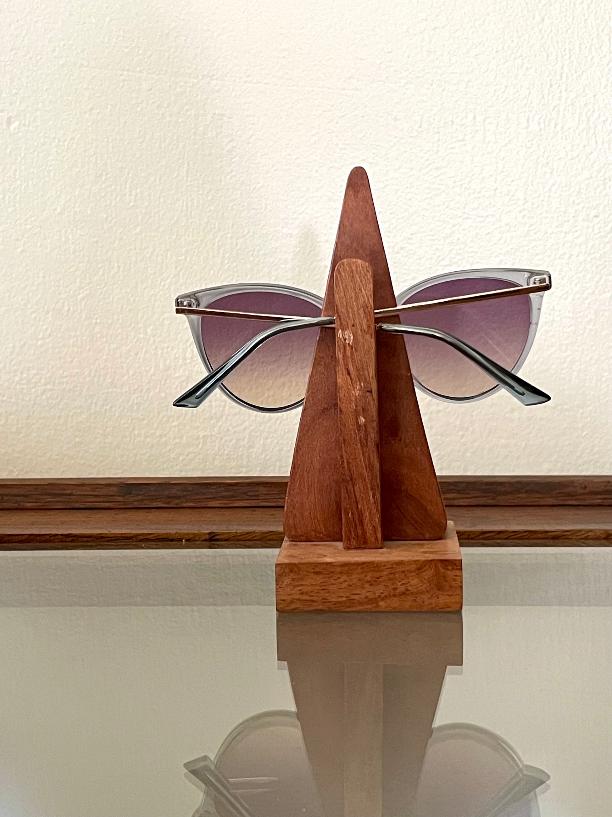 A pair of sunglasses with gradient purple lenses and silver frames shown from the backside as displayed on a triangular wooden stand against a cream-colored wall.