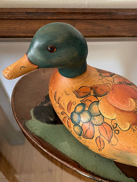 A hand-painted wooden duck decoy figurine with a dark teal-green head, orange beak, and an orange-brown body decorated with painted leaves and grape clusters, resting on an abstract ceramic platter glazed in brown, black, and teal tones.