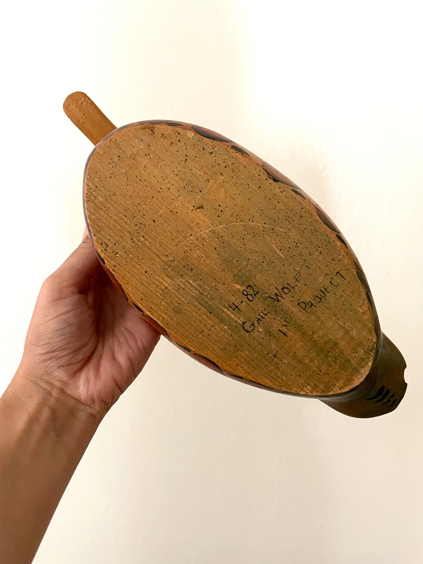 A hand holding an oval wooden object, which is the underside of a wooden duck decoy, against a plain off-white background. Carved into the base is ‘4-82 Gail Wolf 1st Project’.