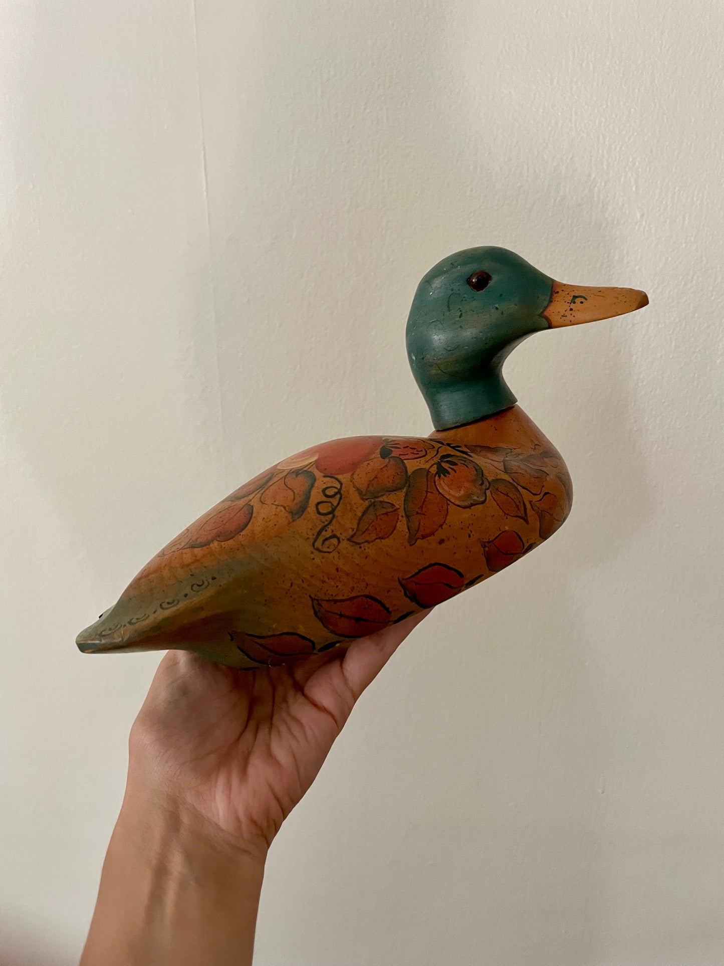 A hand holds a hand-painted wooden duck decoy against a plain off-white wall. The duck has a dark green head, a tan beak, and a brown body decorated with red and orange floral patterns and scrollwork.