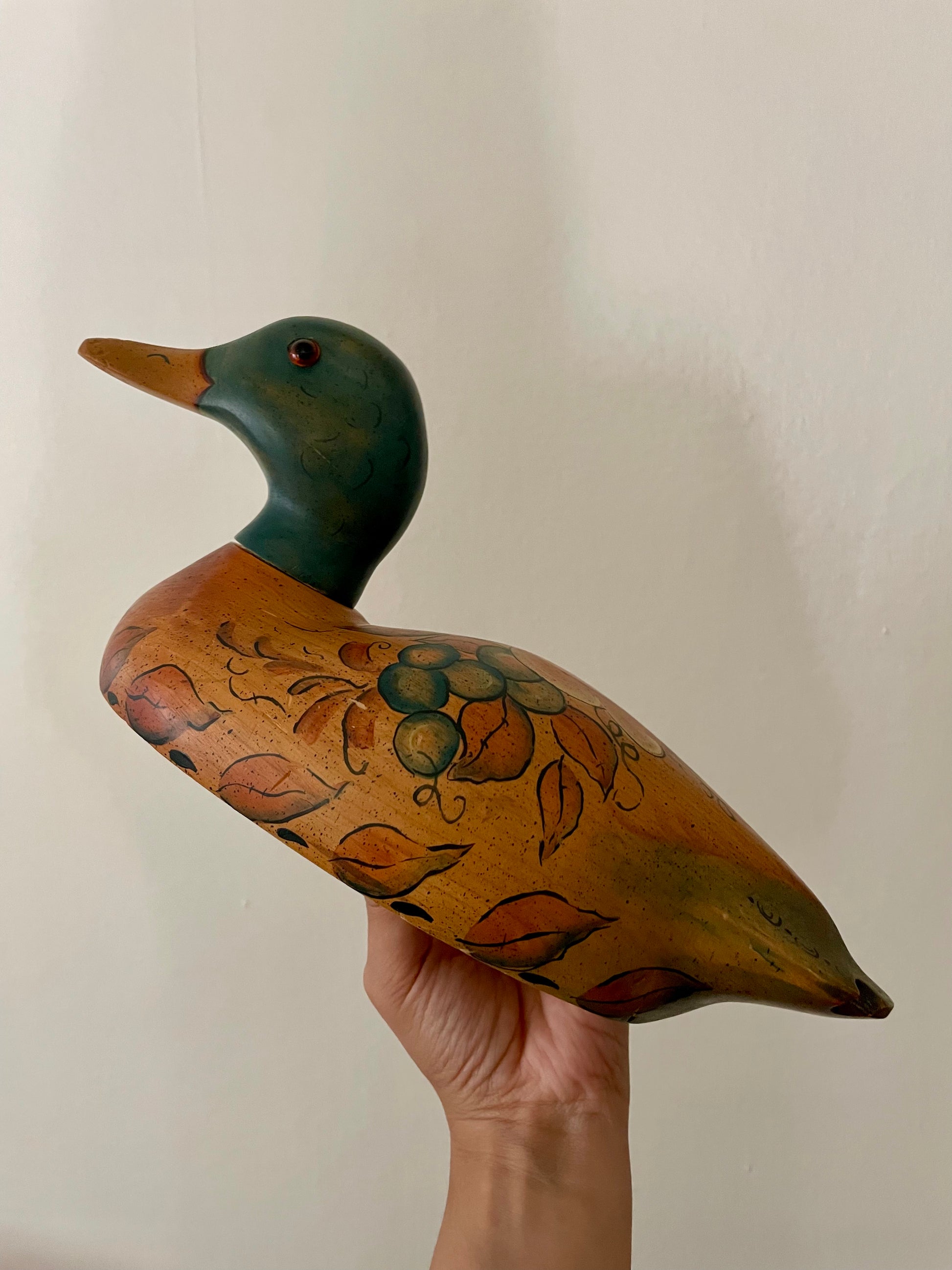 A hand-painted wooden duck decoy held by a person's hand against a plain off-white wall. The duck features a dark green head, a yellow-orange beak, and a warm brown body decorated with painted leaves and berries.
