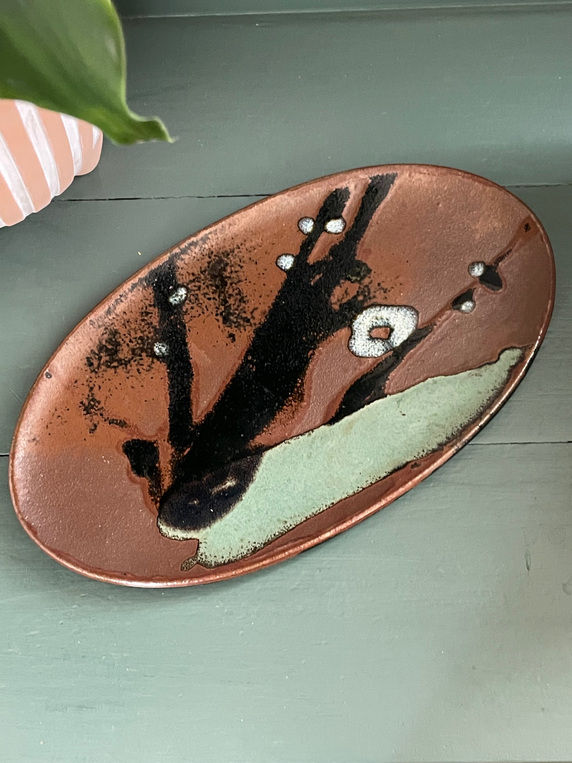 An oval ceramic serving platter with an abstract hand-painted design, featuring a copper-brown base with black brushstrokes, white speckles, and a light sage green section, resting on a muted green wooden surface.