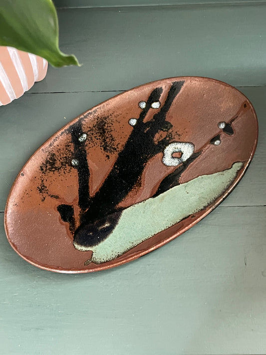 An oval ceramic serving platter with an abstract hand-painted design, featuring a copper-brown base with black brushstrokes, white speckles, and a light sage green section, resting on a muted green wooden surface.