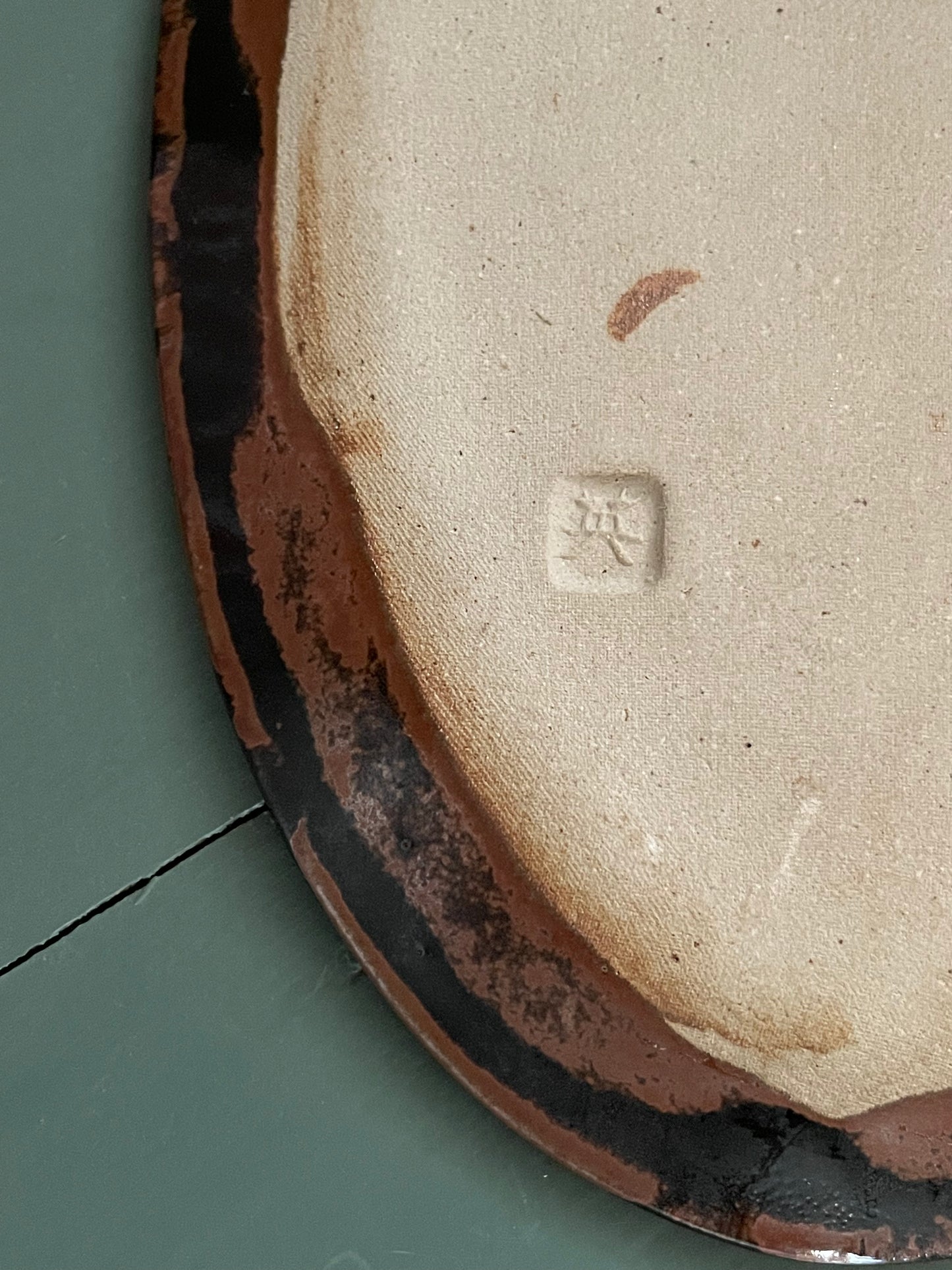 Close-up of the underside of a ceramic  dish, showing a light beige unglazed clay body with a dark brown and black glazed rim on a muted green surface. There is a Japanese maker’s mark stamped into the bottom.