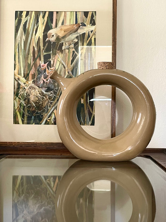 A glossy, abstract ring-shaped decanter in a warm beige/tan color sits on a reflective glass table, with a framed bird painting leaning behind it against a light wall.
