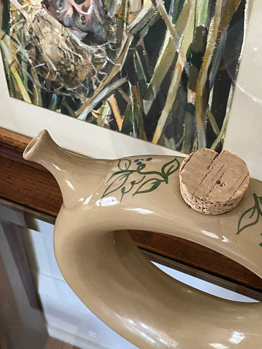 A close-up of a donut-shaped beige ceramic decanter, decorated with green hand-painted leaf patterns and with a cork stopper at top and spout to the left, set against a background of framed artwork and a wooden ledge.
