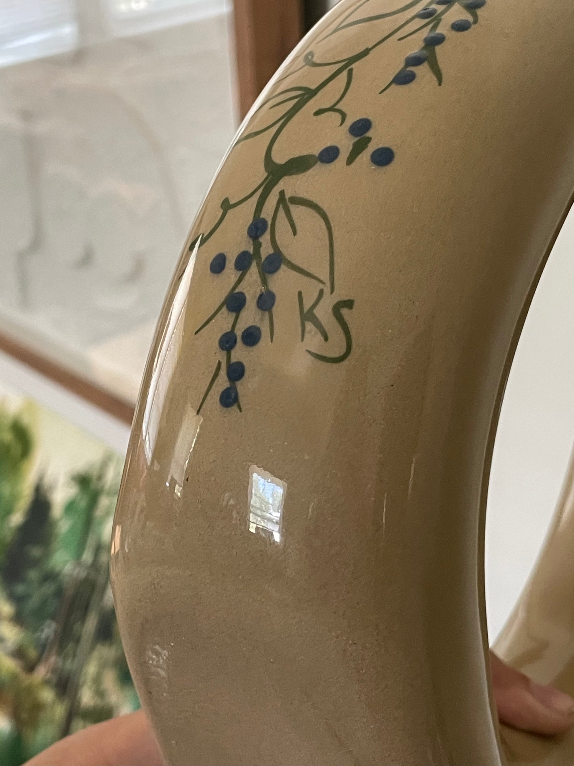 Close-up of the base of a glossy beige donut-shaped ceramic decanter with a hand-painted green and blue berry branch design and signed ‘KS’, held indoors with a blurred background.