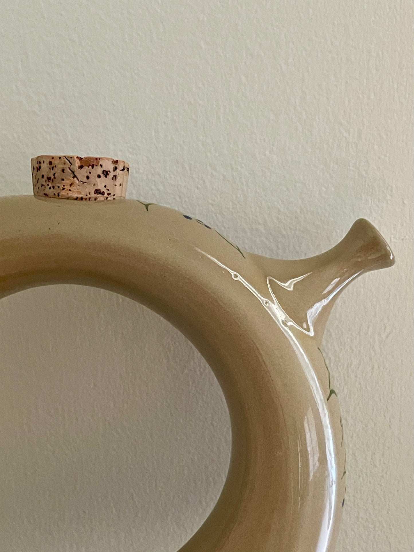 A close-up of a donut-shaped glossy beige ceramic decanter with a small protruding spout and a small speckled cork stopper resting on top, set against a plain off-white wall.