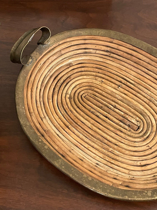 An oval coiled rattan tray with a rustic bronze metal rim and handle, shown on a dark wood background.