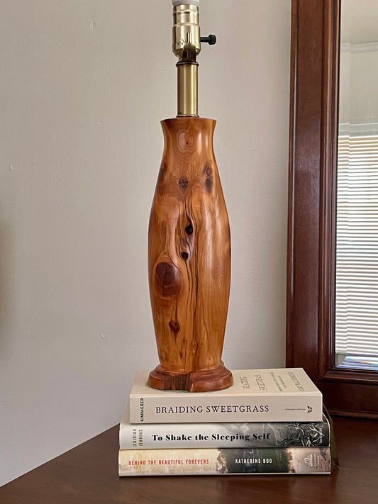 A rustic wooden table lamp with a polished, curved body and a brass-colored neck, displayed on a stack of three books on a wooden table next to a mirror.