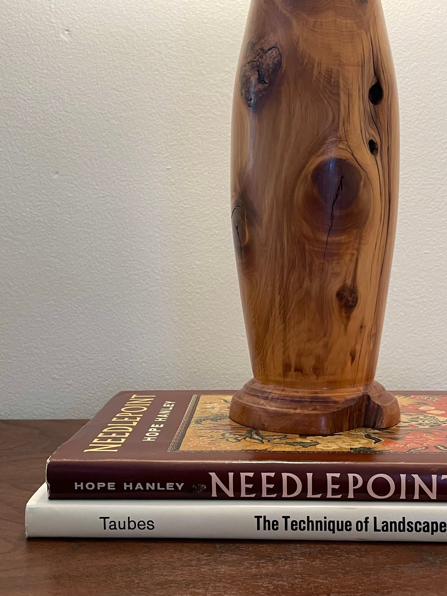 A tall, polished wooden lamp base with natural grain and knot details, displayed on top of two books against a plain wall.