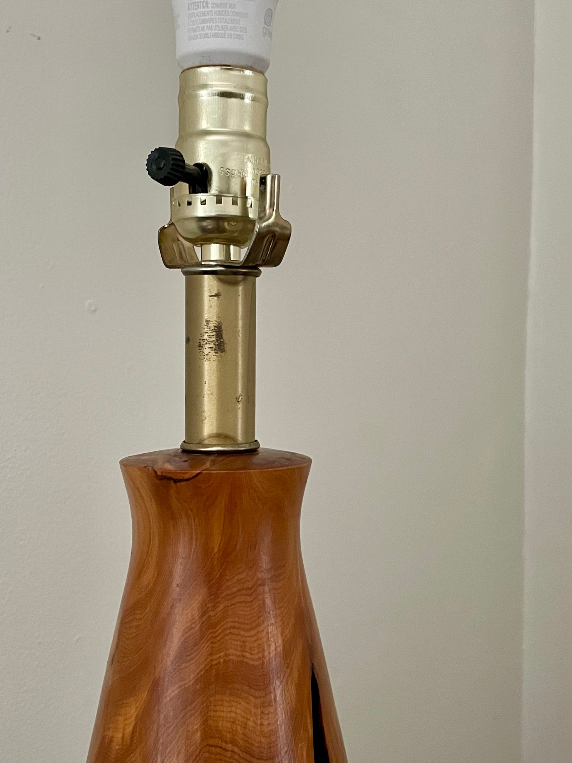 A close-up of a lamp base made from polished wood, featuring a brass-colored metal socket with a black adjustment knob and a white bulb holder at the top, set against a plain beige wall.