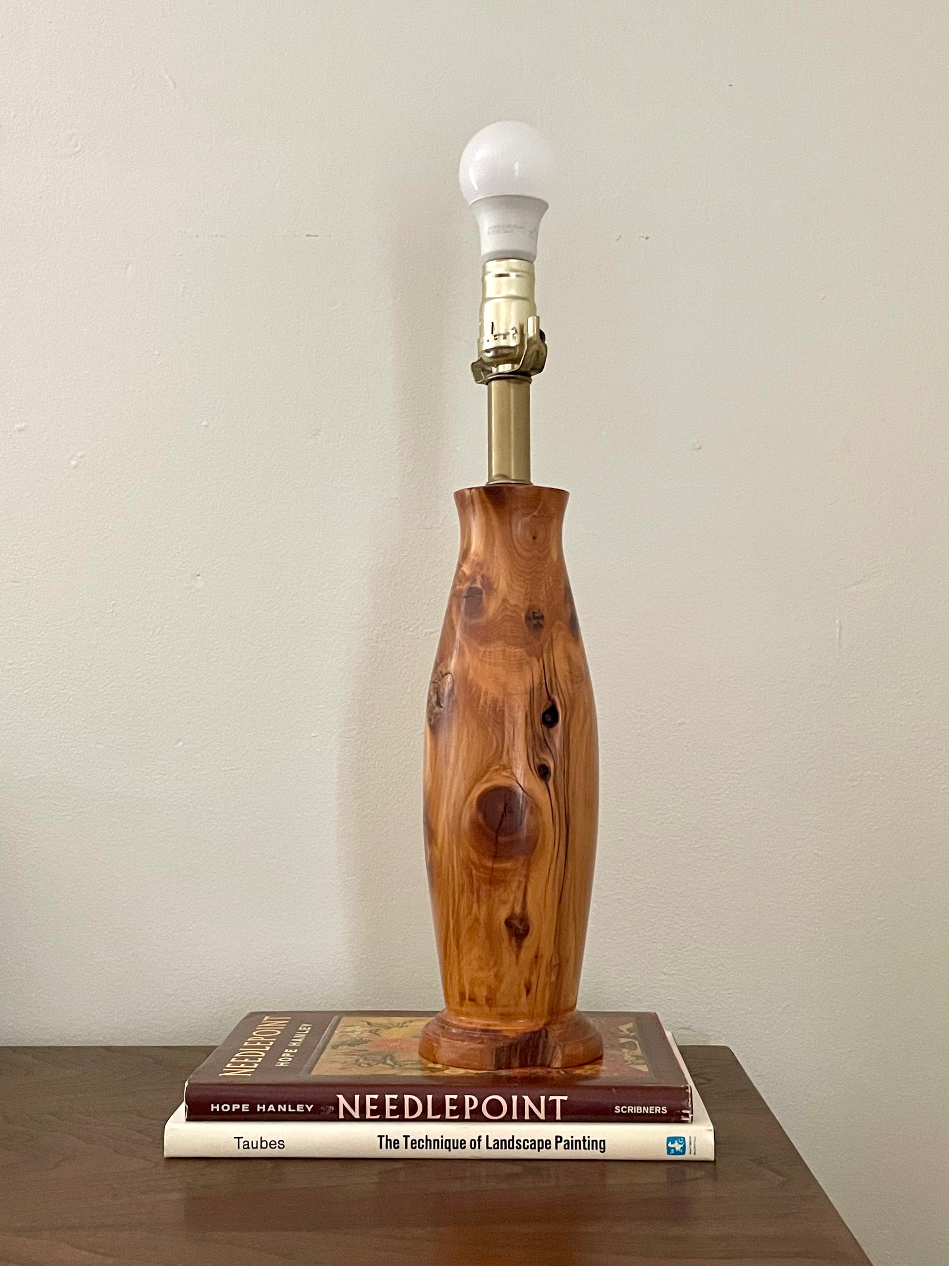 A rustic table lamp with a polished wooden base featuring natural knots and grain, topped with a brass neck and a white bulb, displayed on a stack of books against a plain wall.
