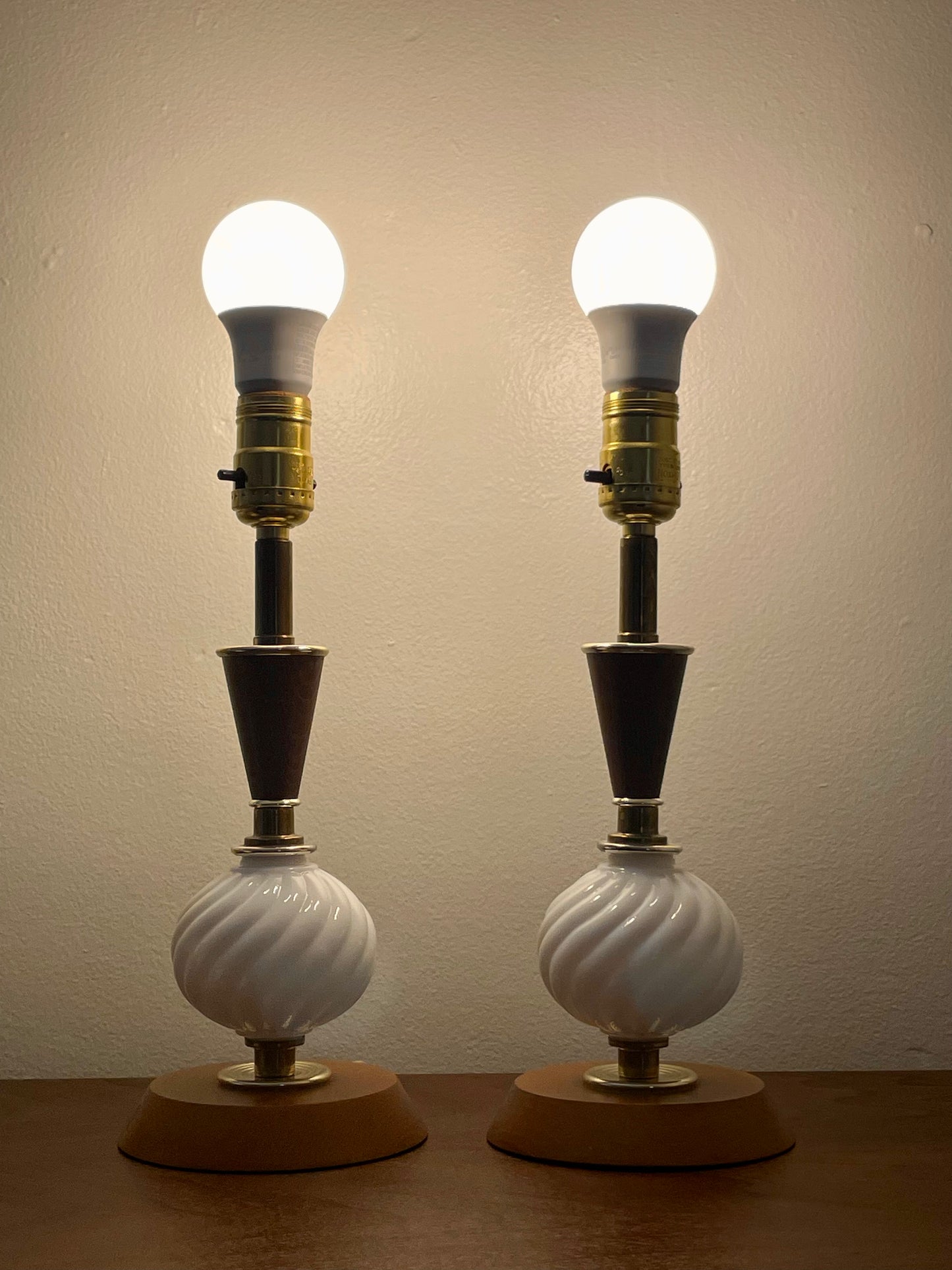 A pair of vintage table lamps with glowing white bulbs, white ribbed glass globes, dark wood neck, brass-toned sockets, and round wooden bases, set against a plain beige wall.