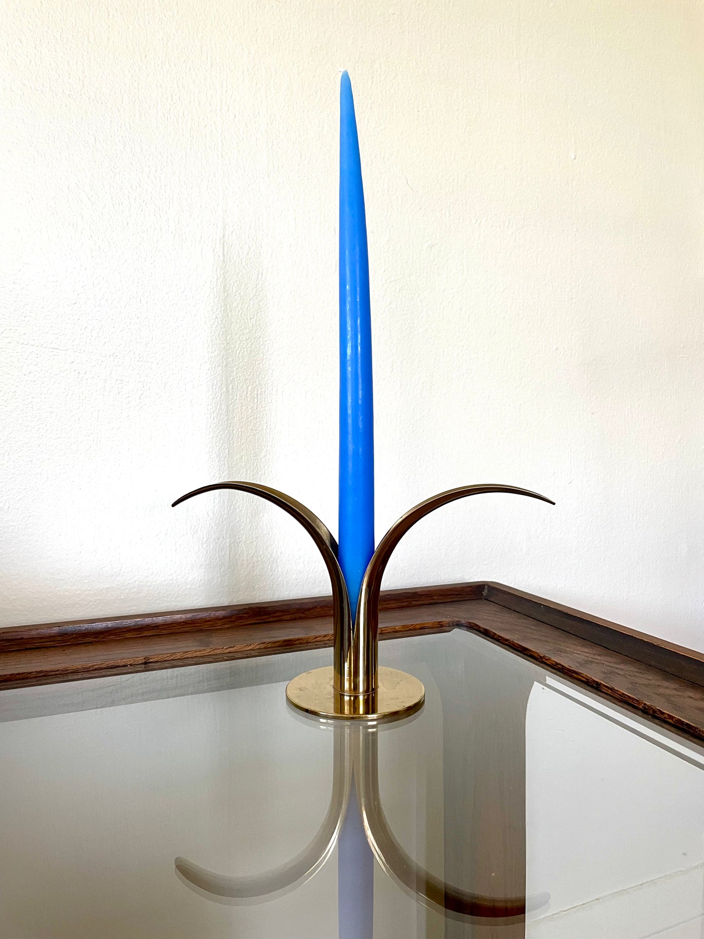 A modern brass candle holder with a tall, slender blue taper candle, displayed on a reflective glass tabletop against a cream-colored wall.