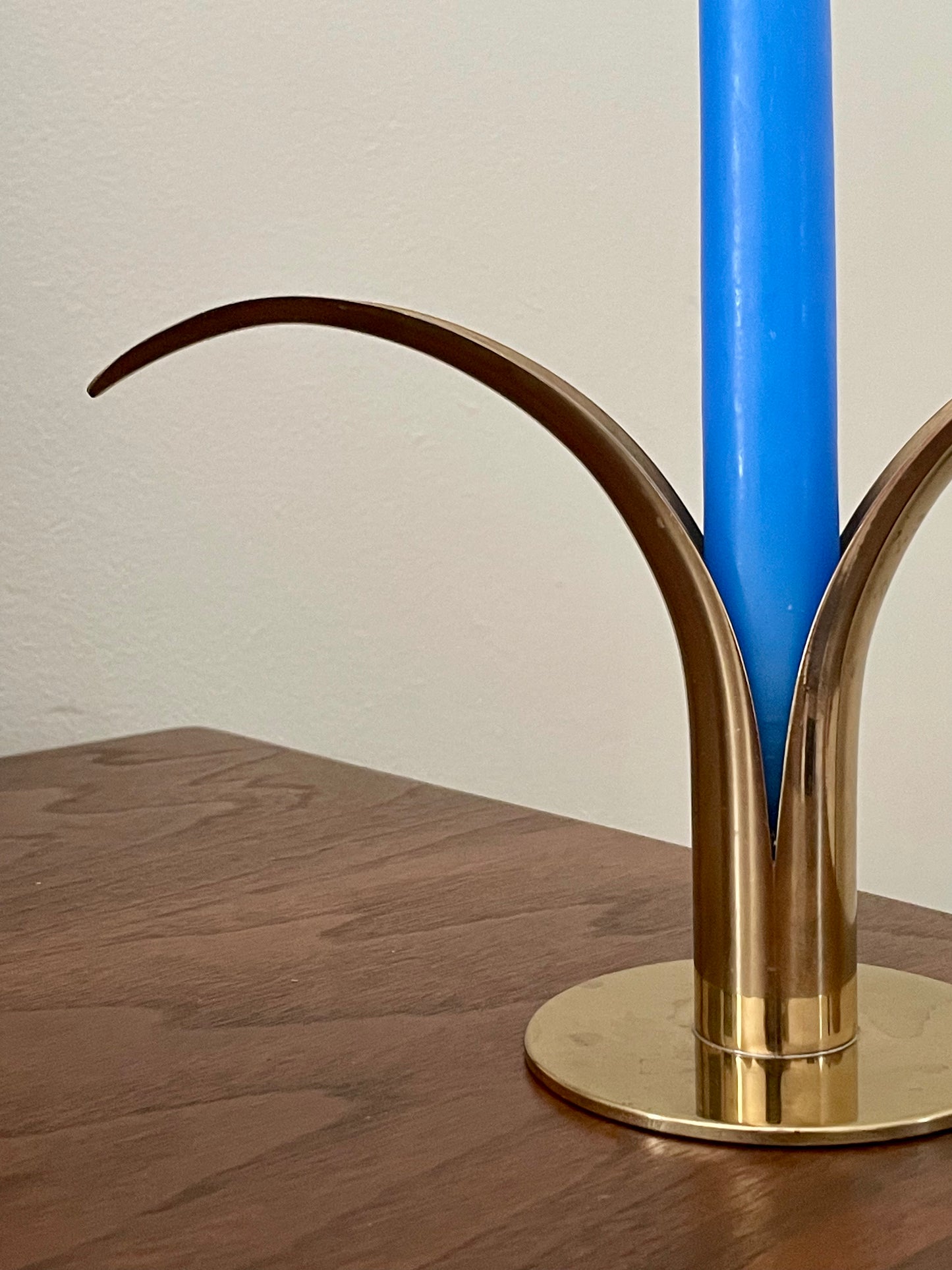 A close-up of a brass sculptural candle holder with curved arms, holding a bright blue candle on a wooden tabletop against a plain off-white wall.