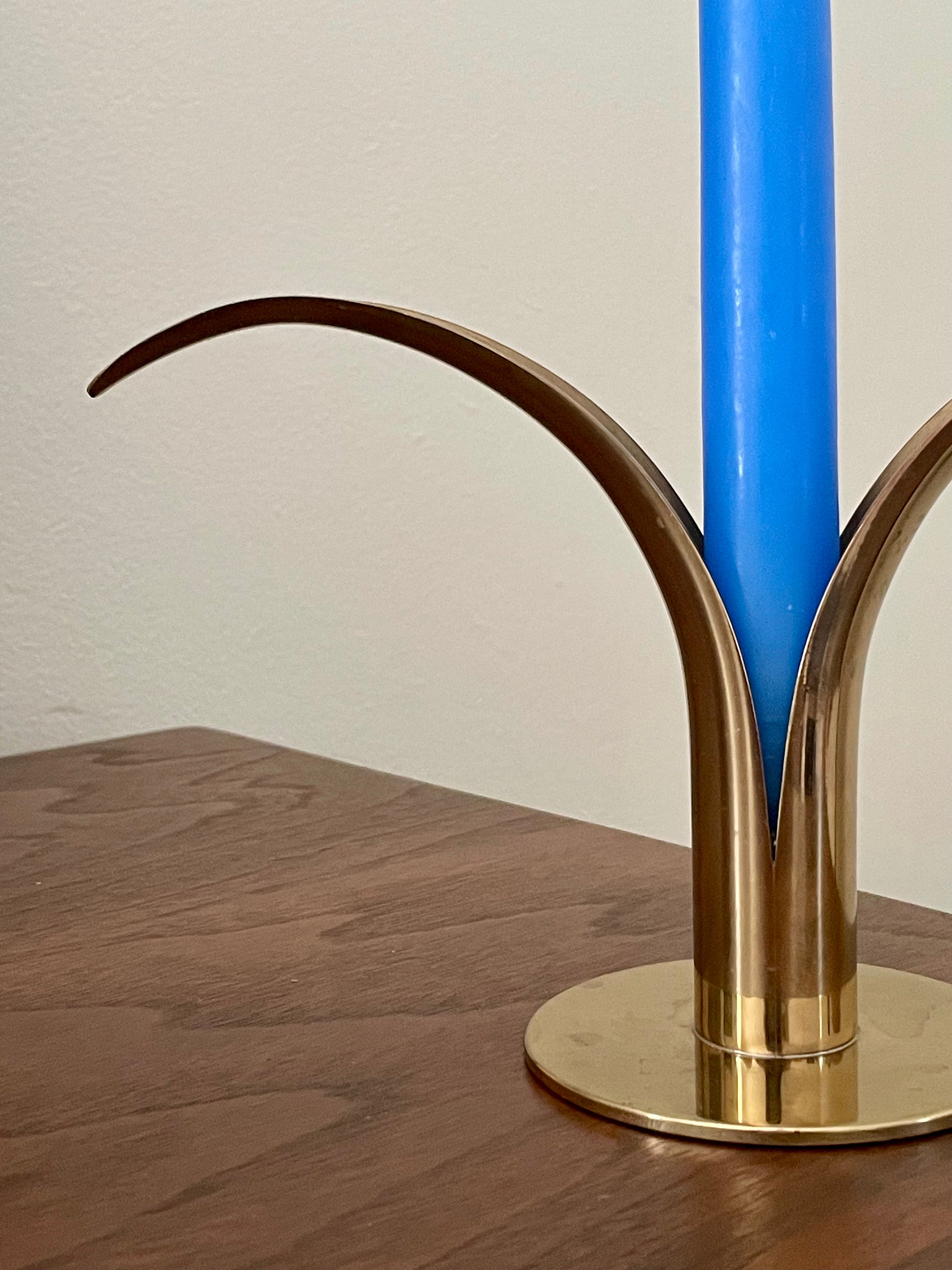 A close-up of a brass sculptural candle holder with curved arms, holding a bright blue candle on a wooden tabletop against a plain off-white wall.