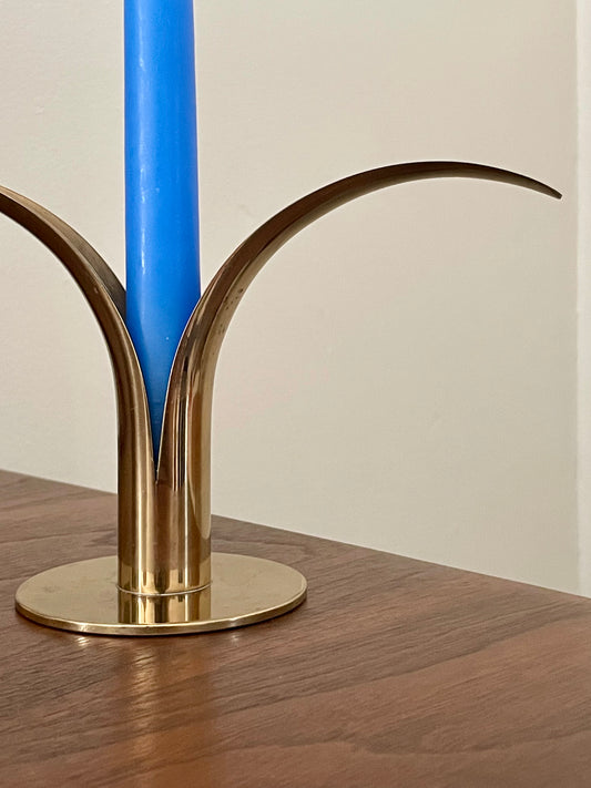 A close-up of a polished brass candlestick holder with curved leaf-like arms, holding a bright blue candle on a wooden tabletop against a plain light wall.