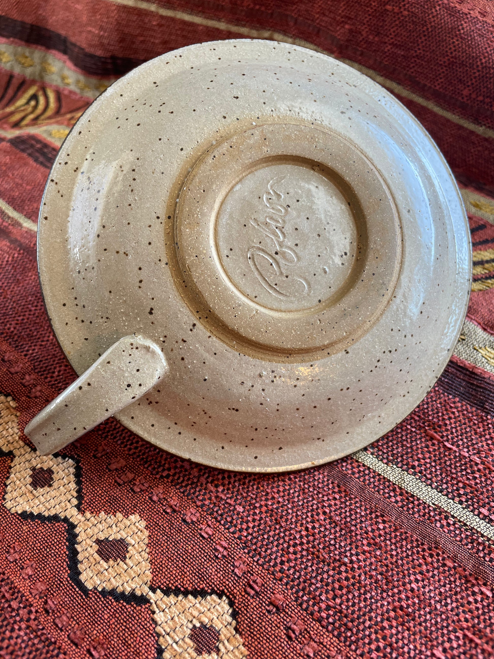 View of the underside of a circular ceramic candle holder with handle resting on a patterned fabric background.