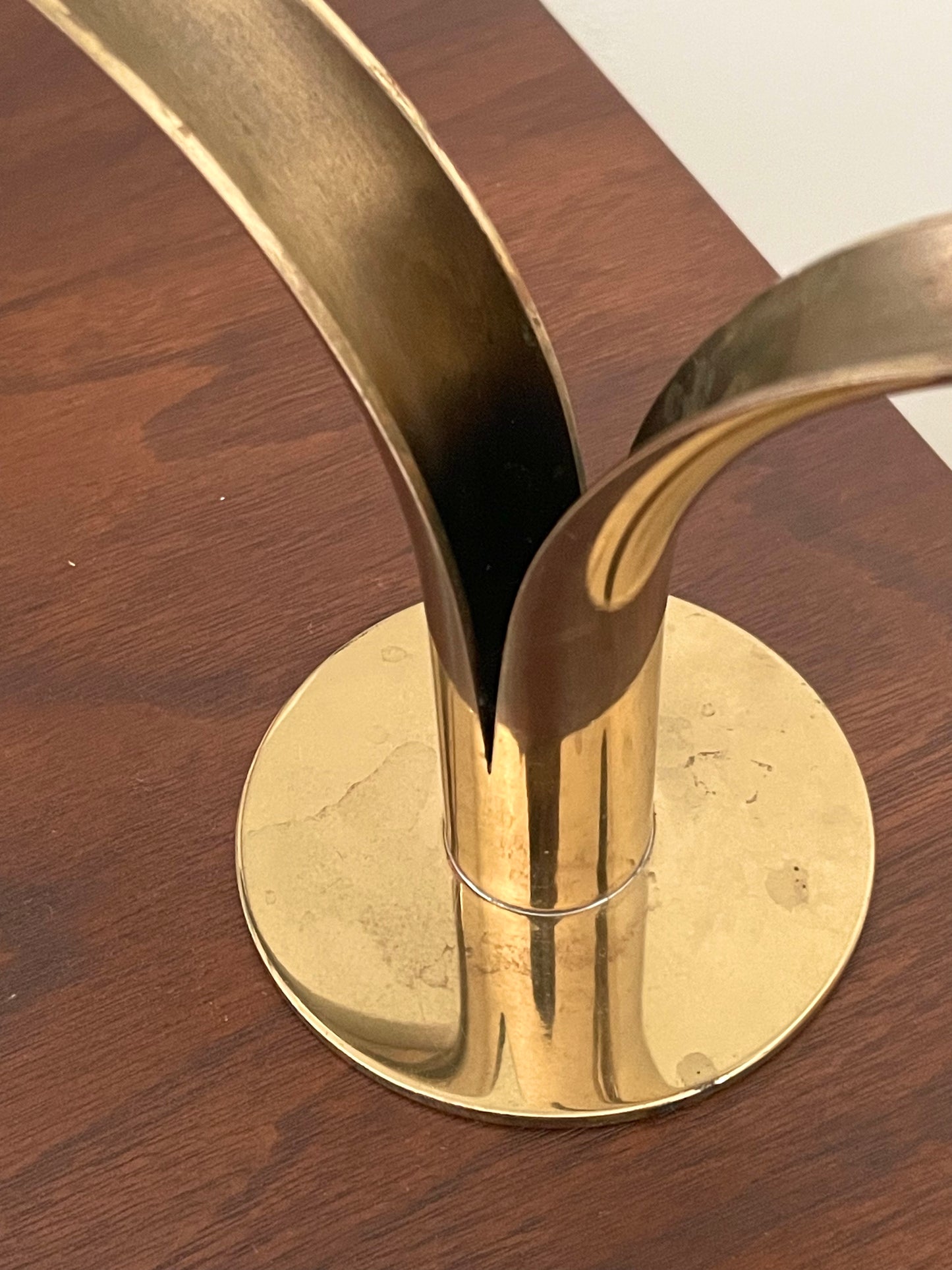A close-up of a polished brass candleholder with a circular base and two upward-curving arms, resting on a dark wood surface.