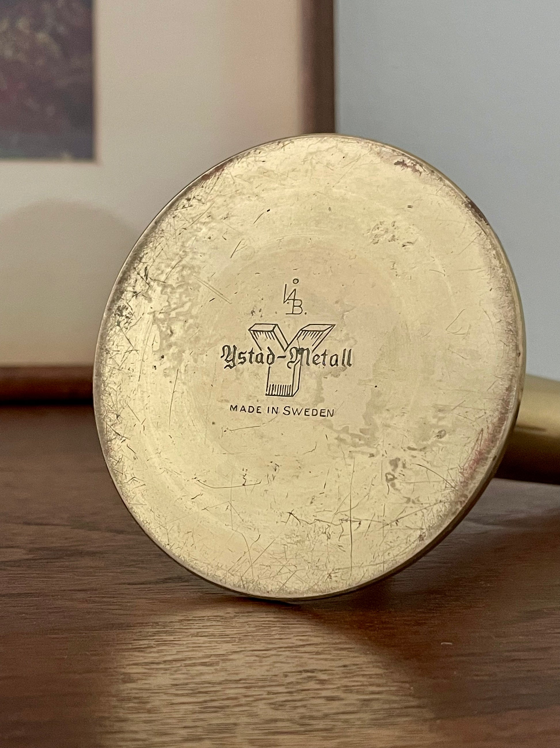 A round vintage brass candleholder lays on its side with the base facing outward, with engraved text reading 'Ystad-Metall' and 'MADE IN SWEDEN', resting on a wooden surface.