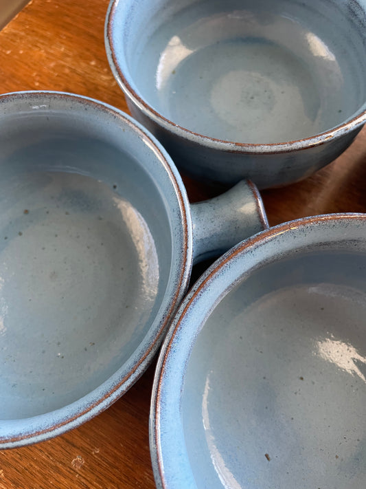Three ceramic bowls glazed in light blue with hints of the underlying terracotta clay peeking through at the rim, arranged on a wooden surface.