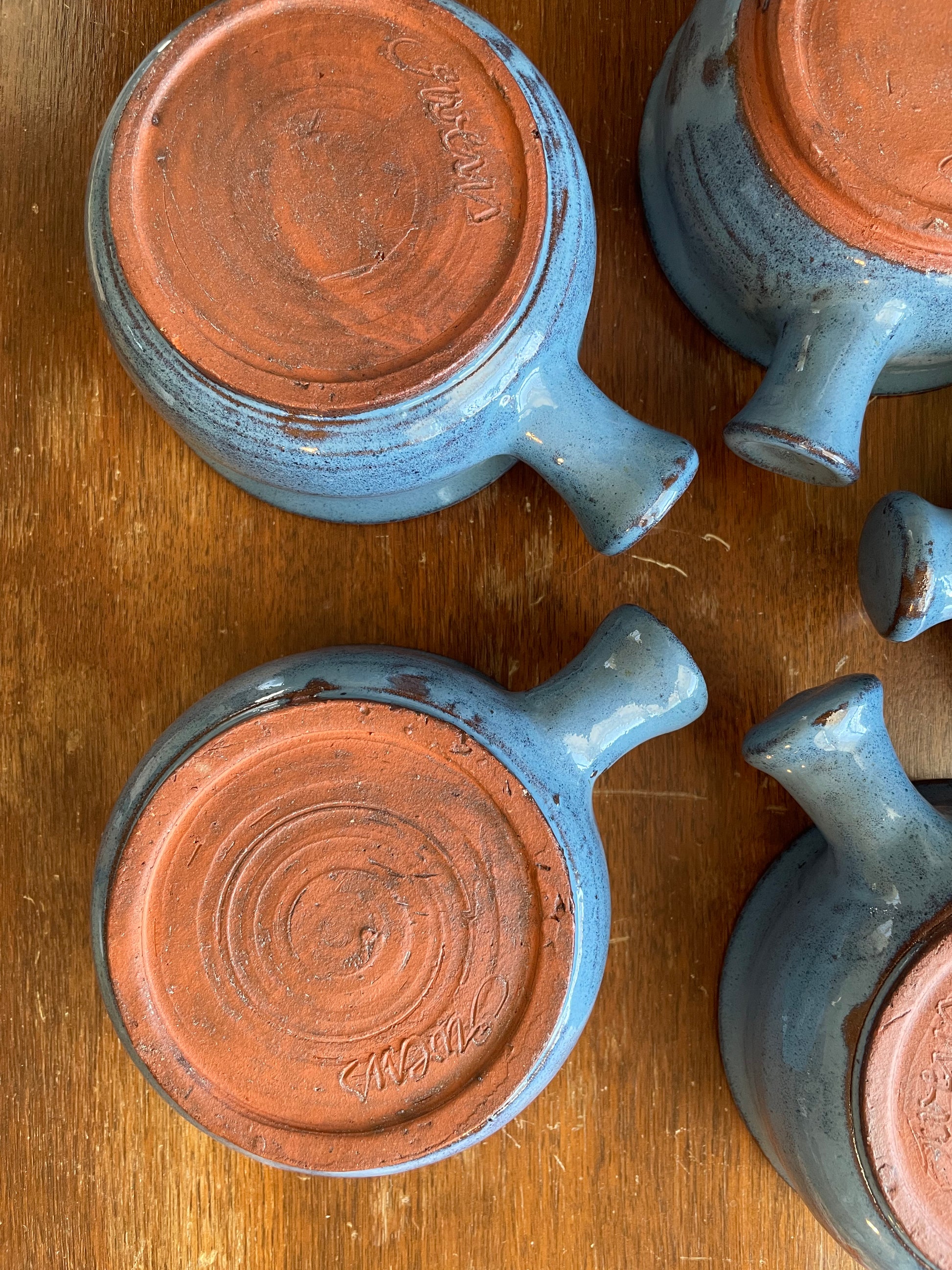 A close-up overhead view of five handmade handled pottery bowls laying upside down and arranged in a circle on a wooden surface. The bowls have a light blue-gray glaze and an unglazed terracotta clay base, and are signed ‘Owens' on the bottom.