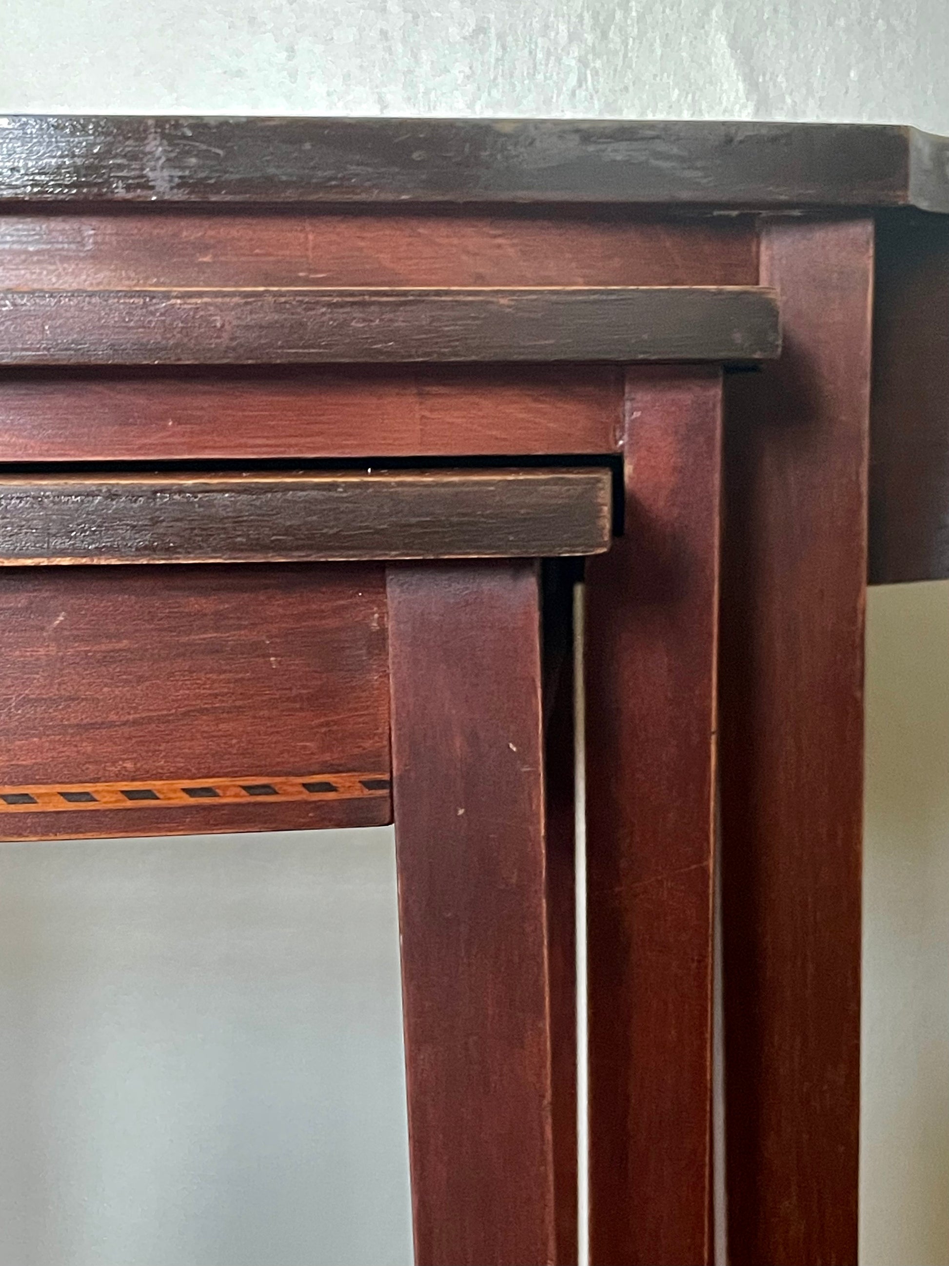 Close-up of a dark reddish-brown set of three nesting tables showing the front tabletop edge, legs, and a small geometric inlay detail against a plain light background.