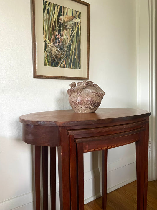A dark reddish-brown wooden console table with a curved top and two nesting tables beneath, positioned against a white wall under a framed bird painting. A textured, earth-toned ceramic vase with lizard accents sits on the table.
