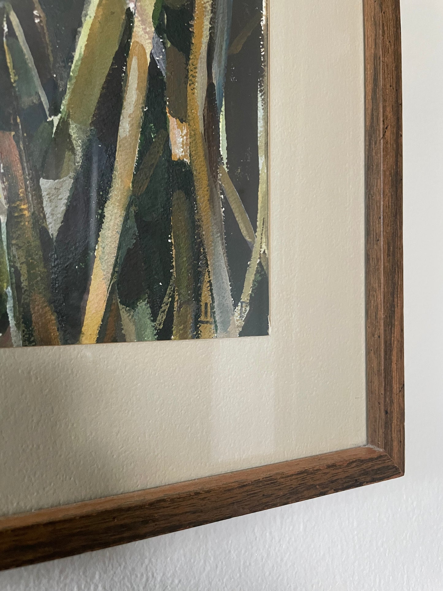 Close-up of a framed painting showing a dark wood frame, cream mat, and painted brushstrokes in black, olive green, beige, and gold tones set against a white wall.