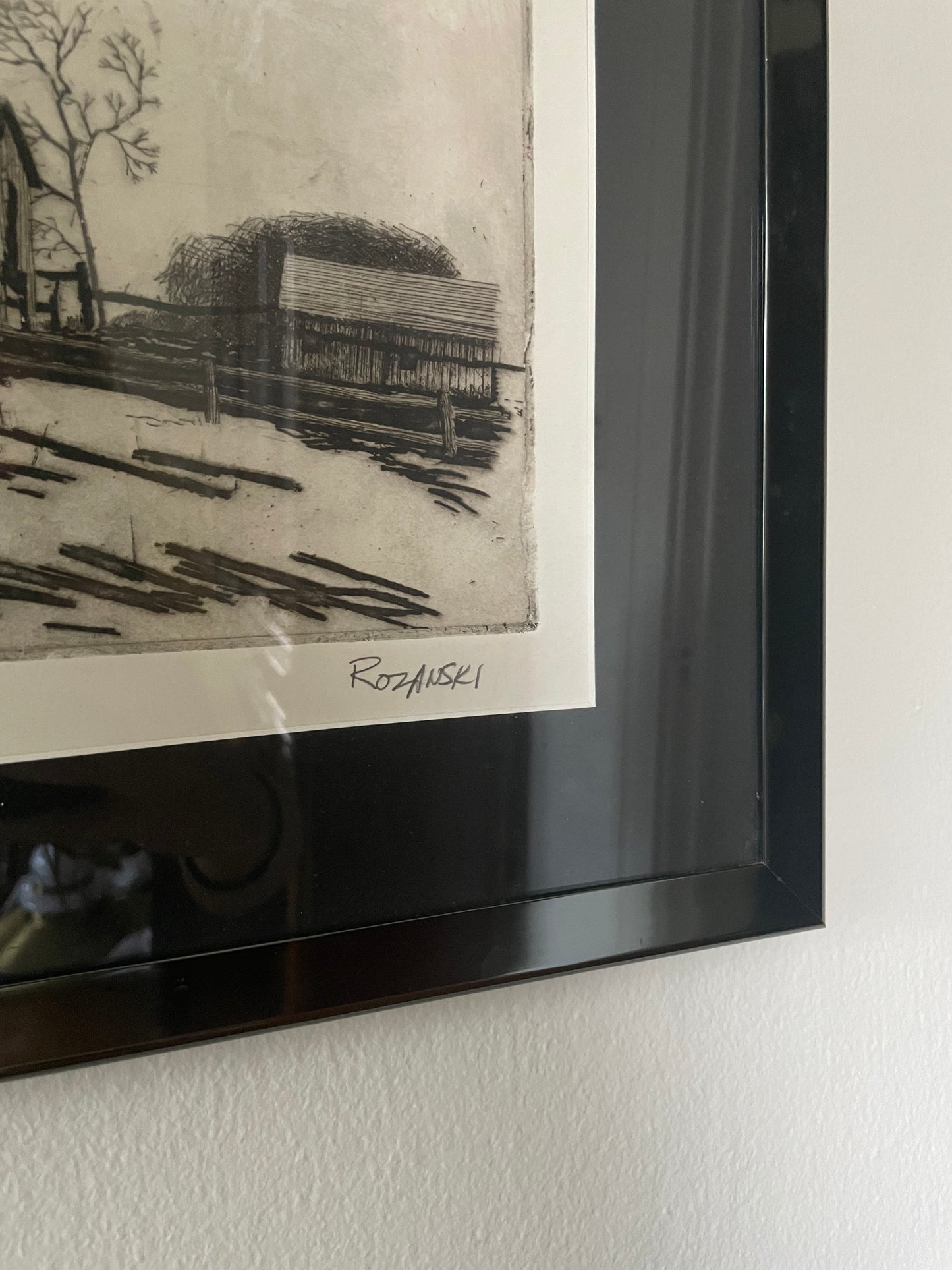 A framed monochrome art print hanging on a white wall, showing a black-and-white print signed ‘Rozanski’ near the bottom and in a glossy black frame.