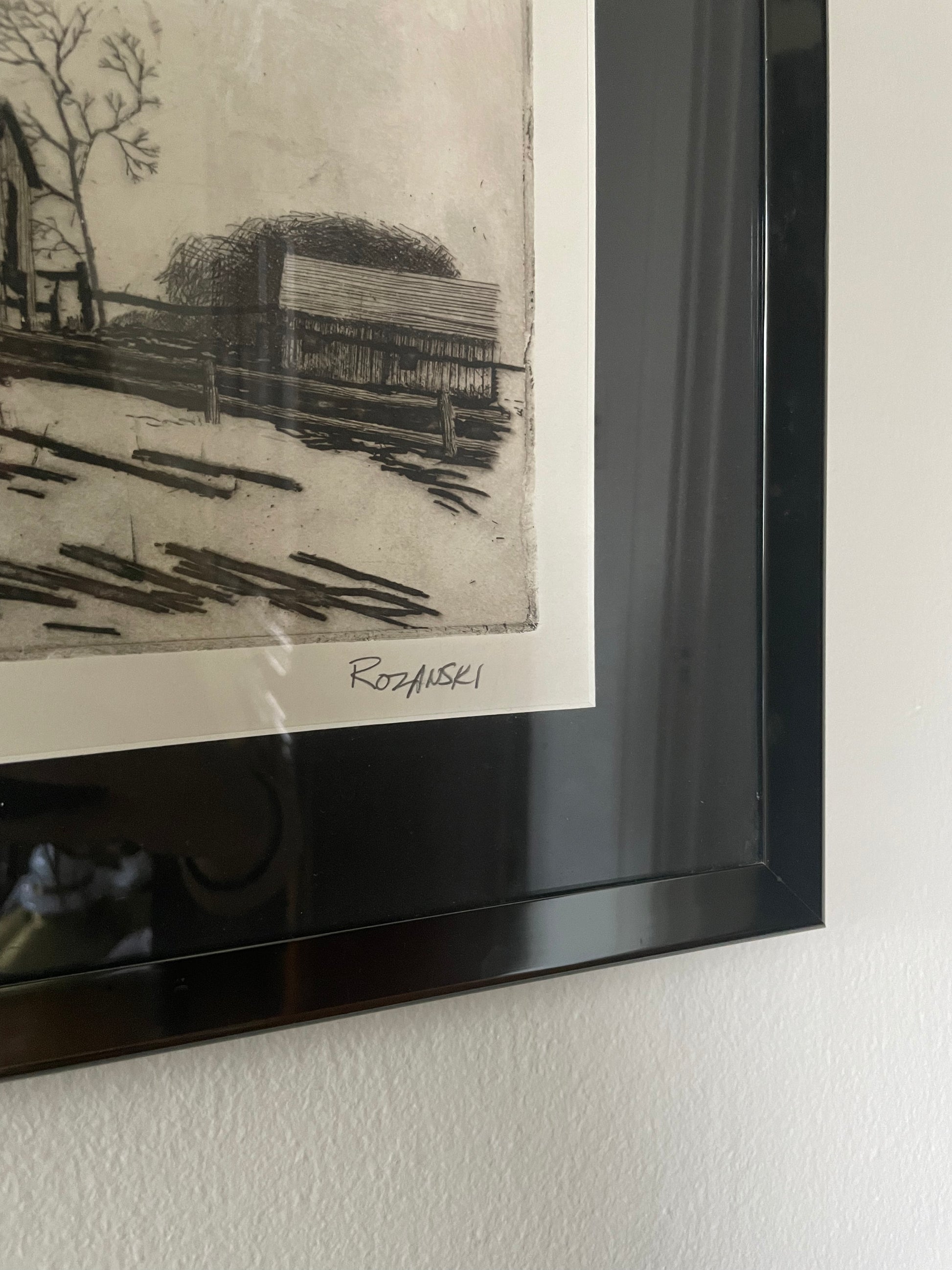 A framed monochrome art print hanging on a white wall, showing a black-and-white print signed ‘Rozanski’ near the bottom and in a glossy black frame.