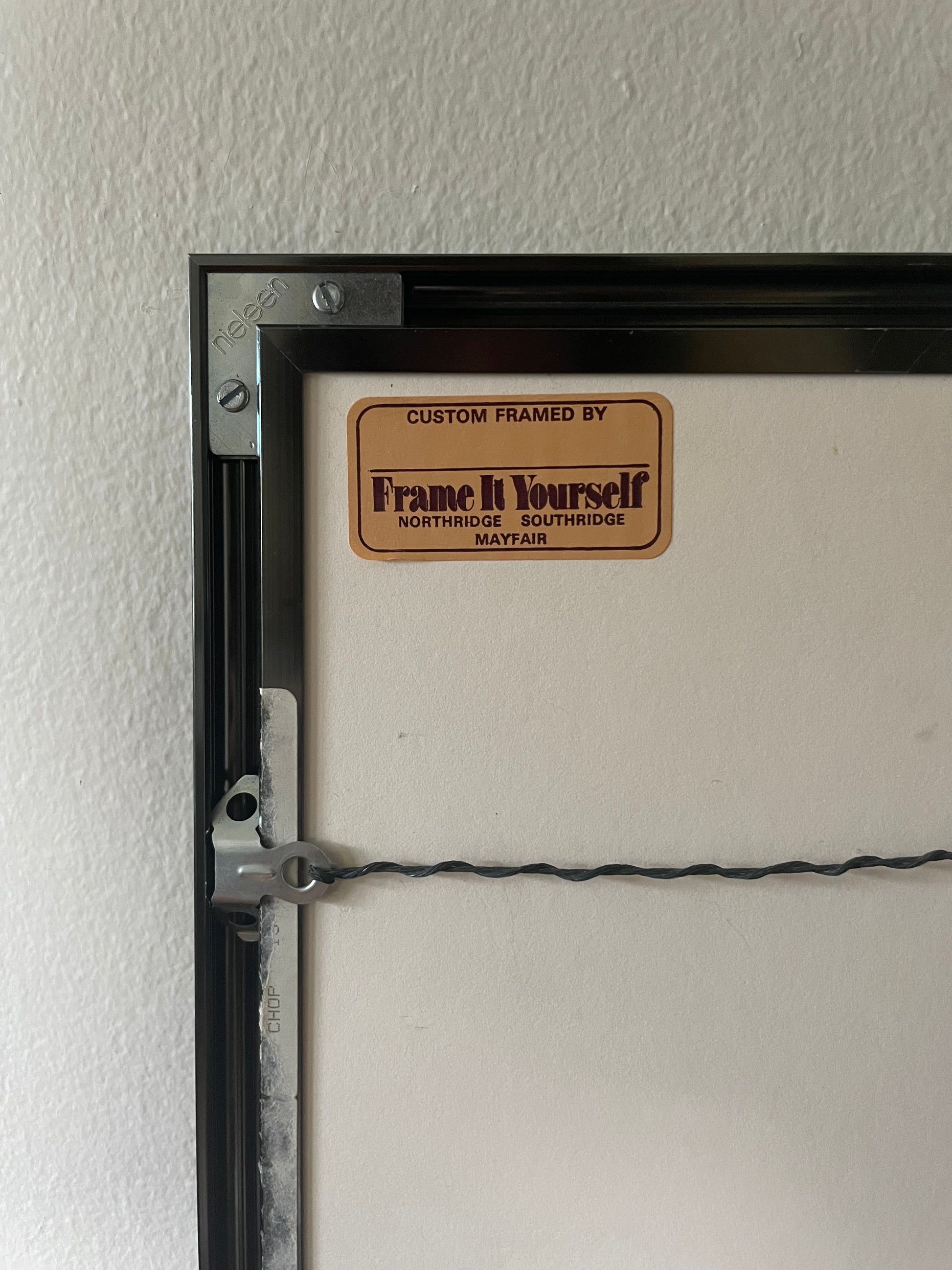Close-up of the back side of a custom framed print, showing a black frame edge, mounting hardware, a wire support, and a tan label that reads 'Custom Framed by Frame It Yourself' on a textured off-white wall.