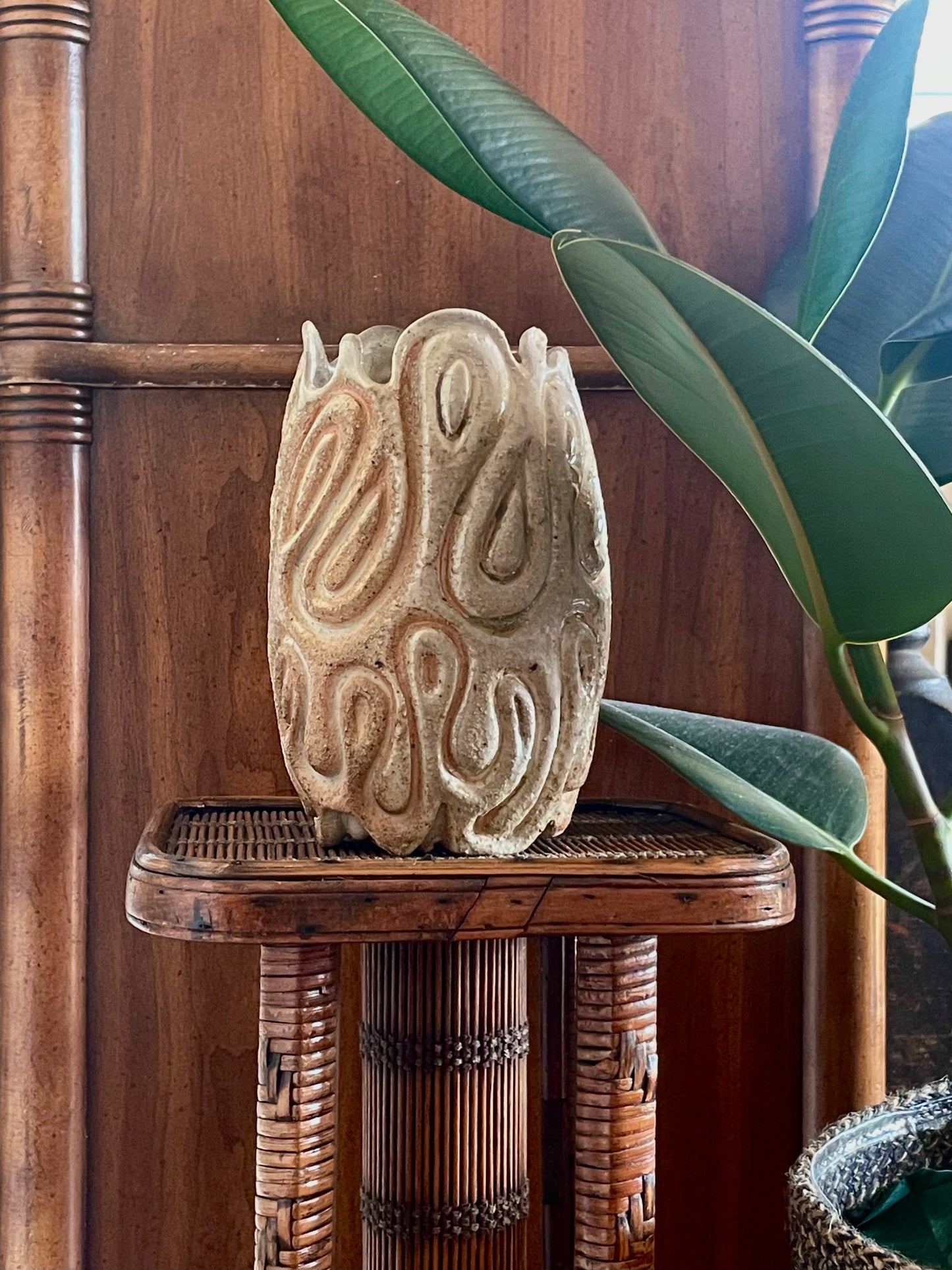 A carved ceramic sculpture in toasty beige glaze with organic, swirling patterns, displayed on a small woven rattan stand next to a green plant in front of a wooden background.